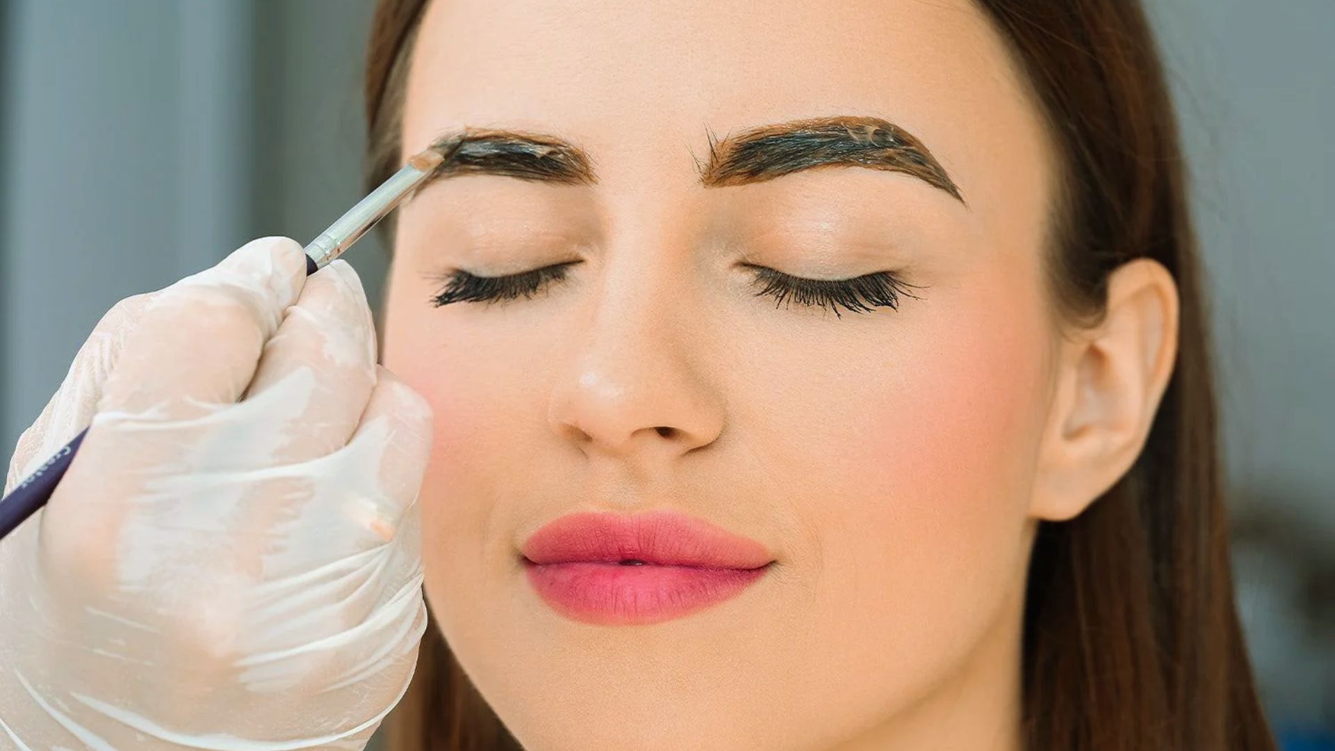 A woman sitting in a salon chair while a beautician shapes her eyebrows with tweezers.
