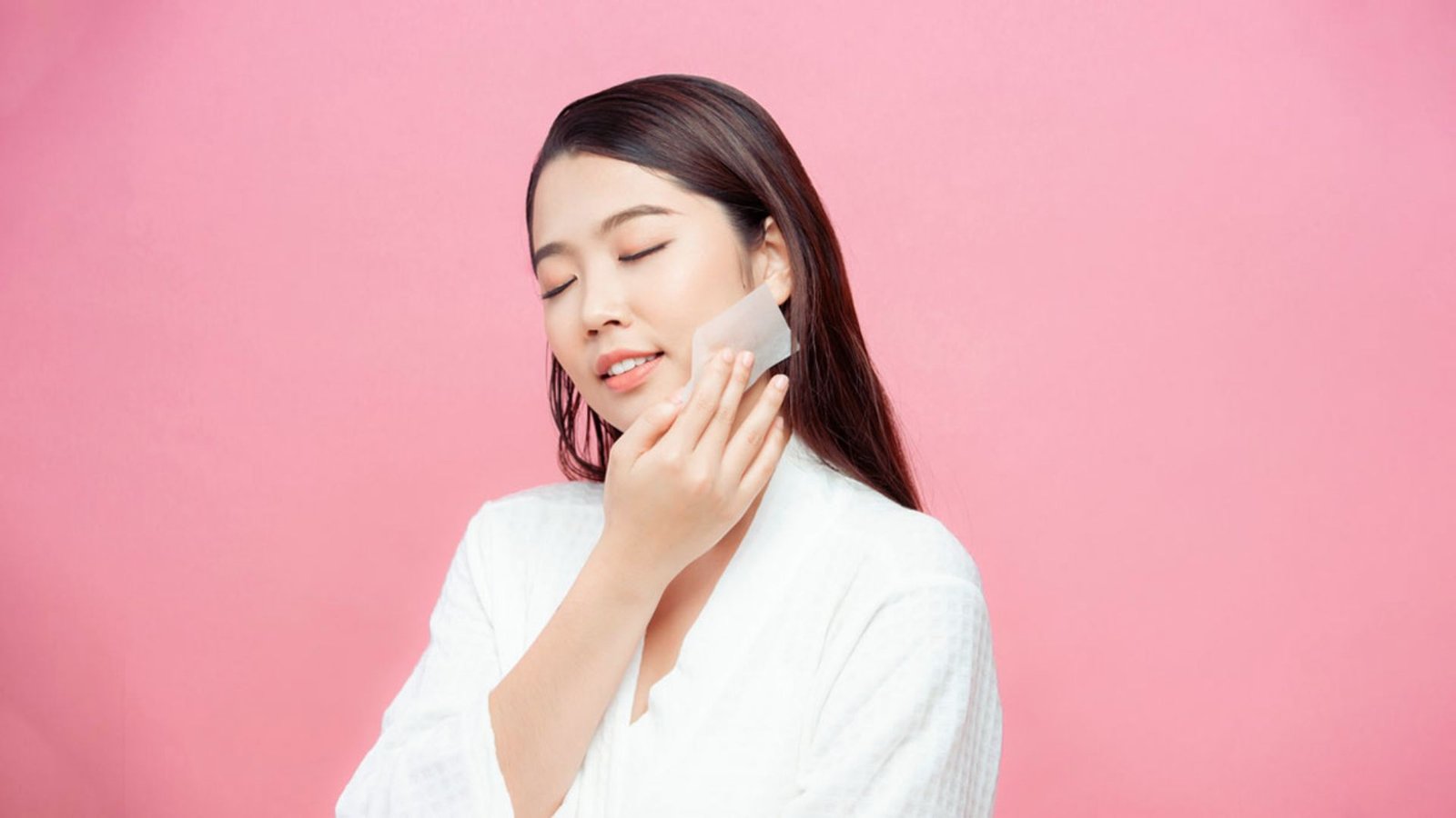 Asian woman in a white robe gently applying a sponge to her face, focusing on skincare.
