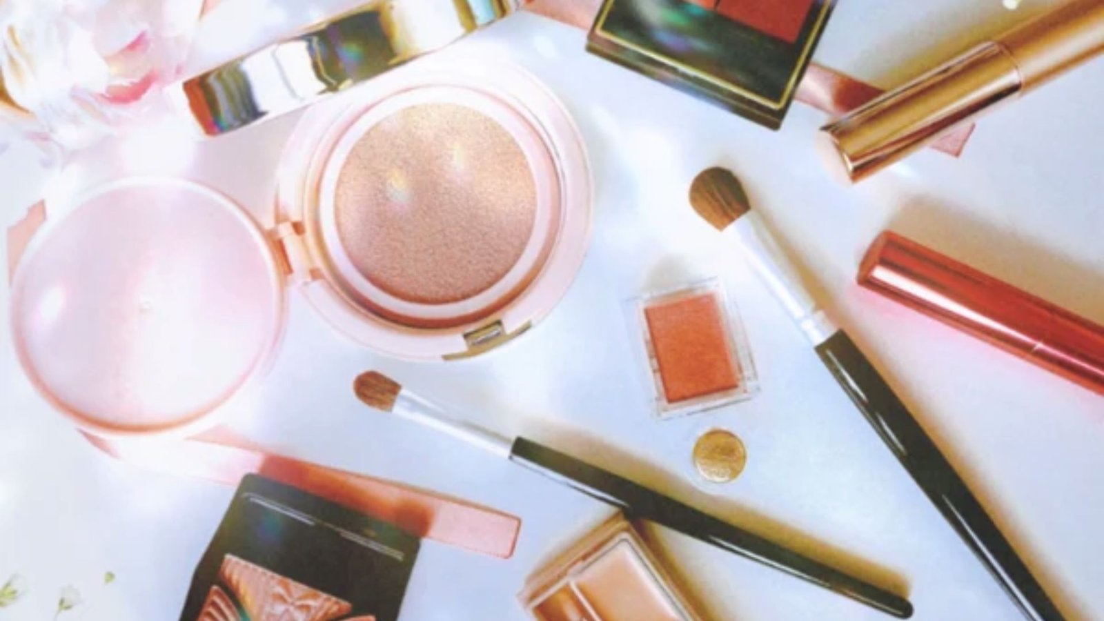 A collection of makeup products arranged on a table beside a delicate pink rose.