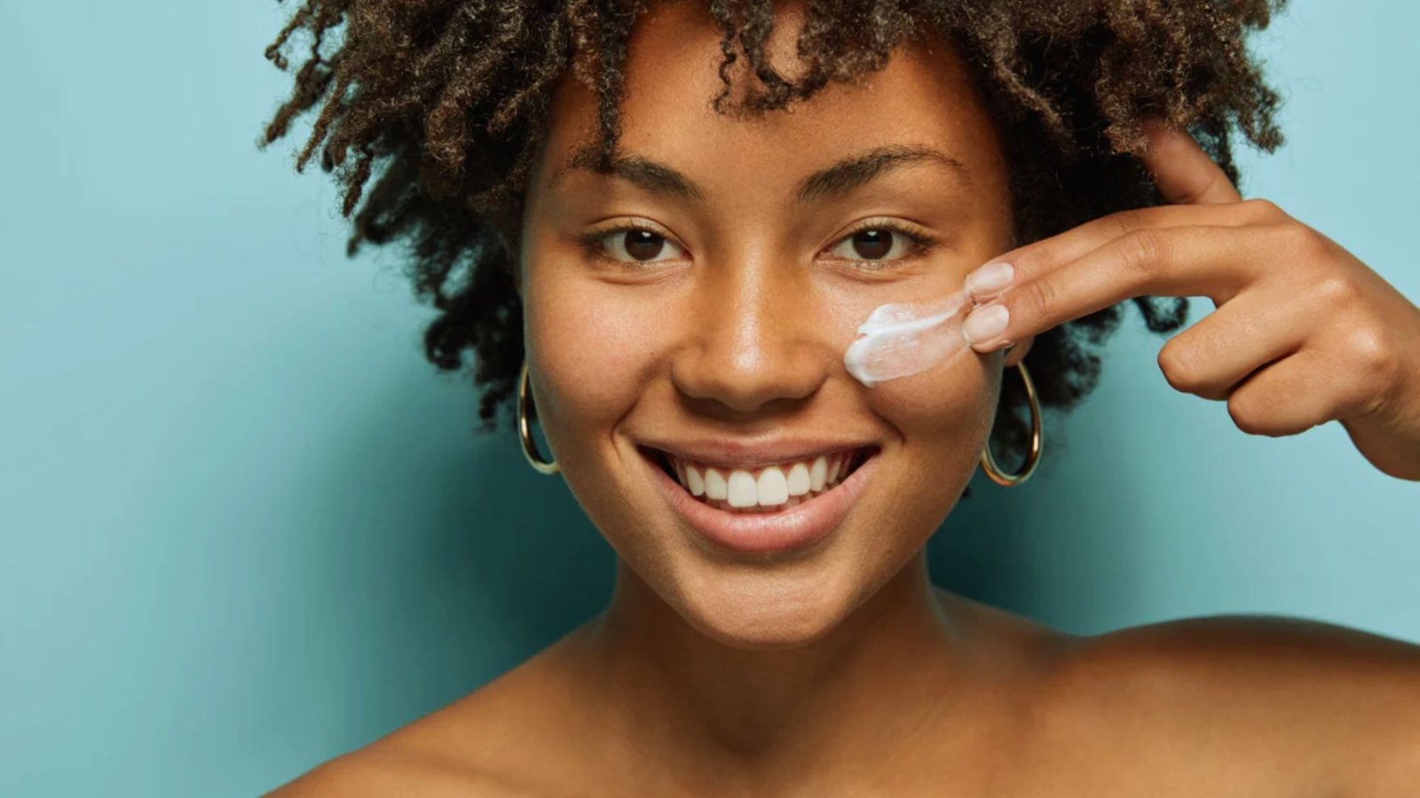 A woman with curly hair smiles while holding a cream, exuding happiness and positivity.