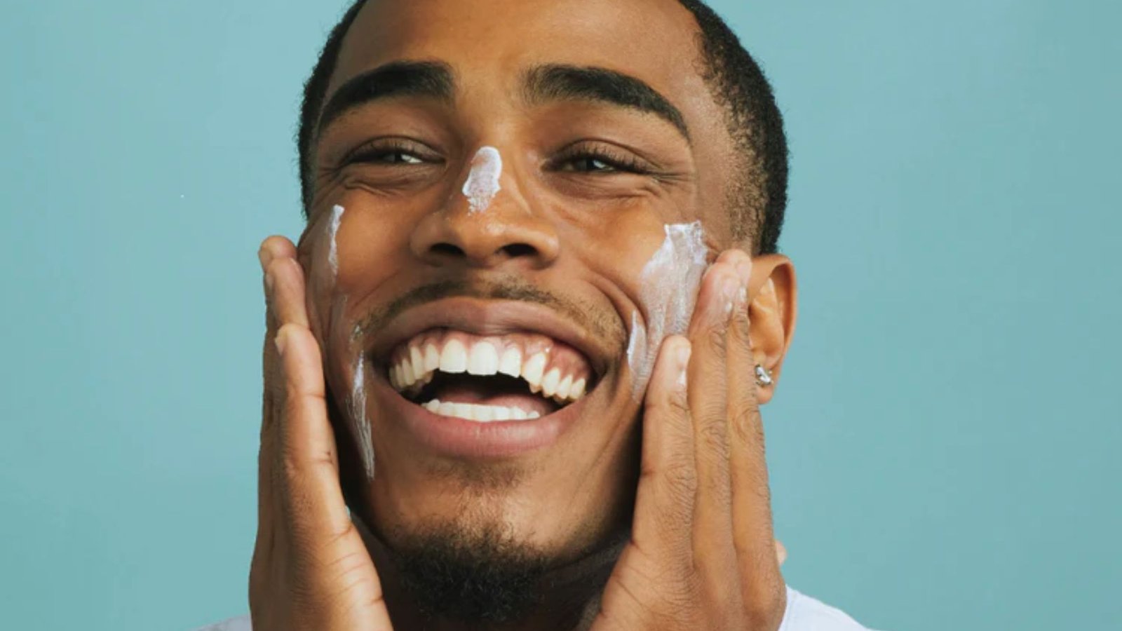 A smiling man applies cream to his face, showcasing a moment of self-care and grooming.