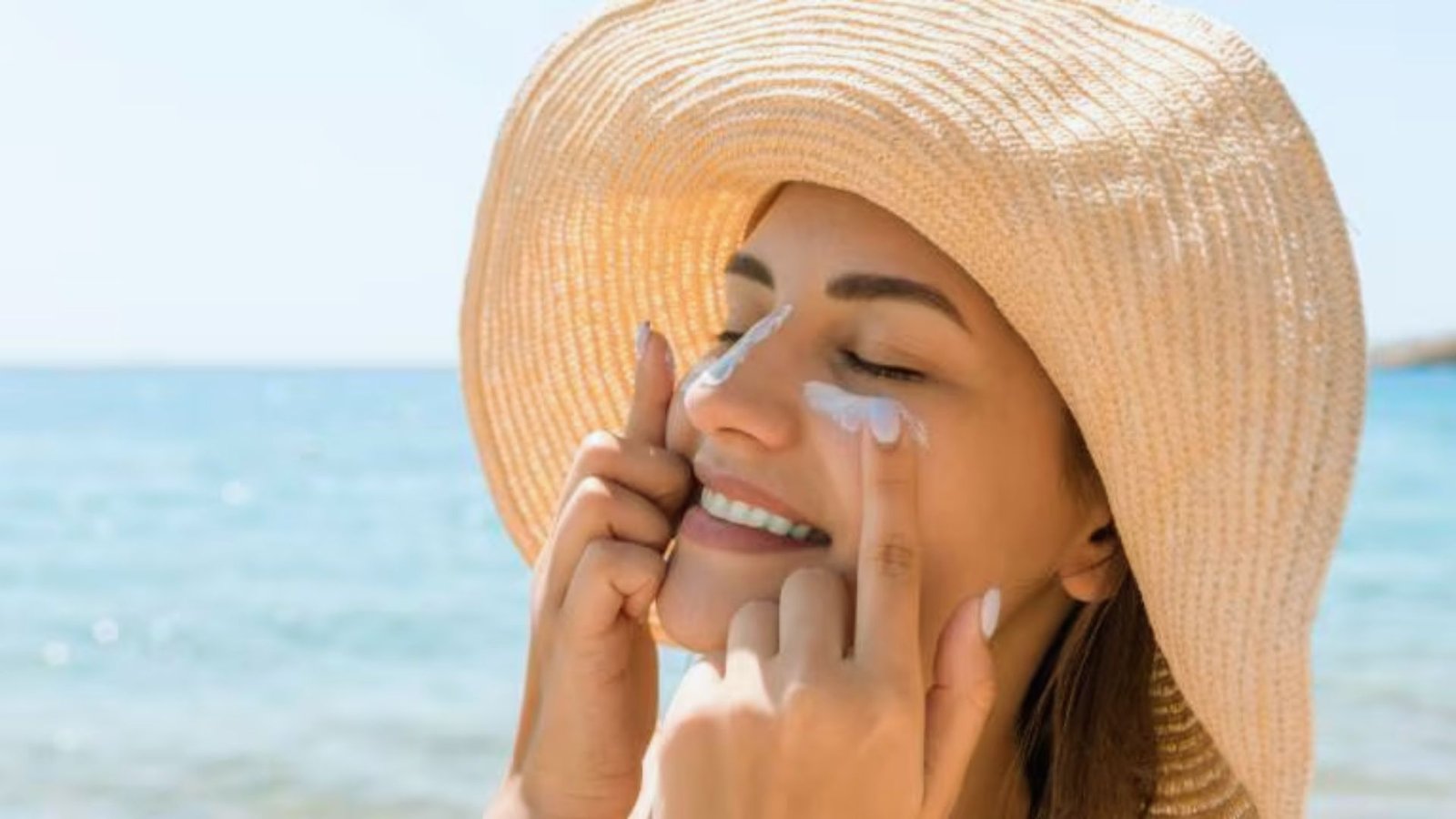 A woman wearing a hat applies sunscreen to her face, preparing for sun exposure.
