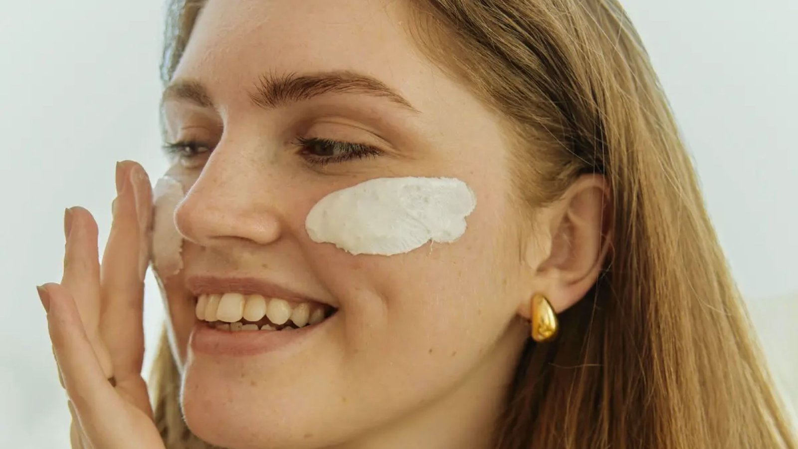 A woman smiles as she applies cream to her face, showcasing a moment of self-care and beauty routine.