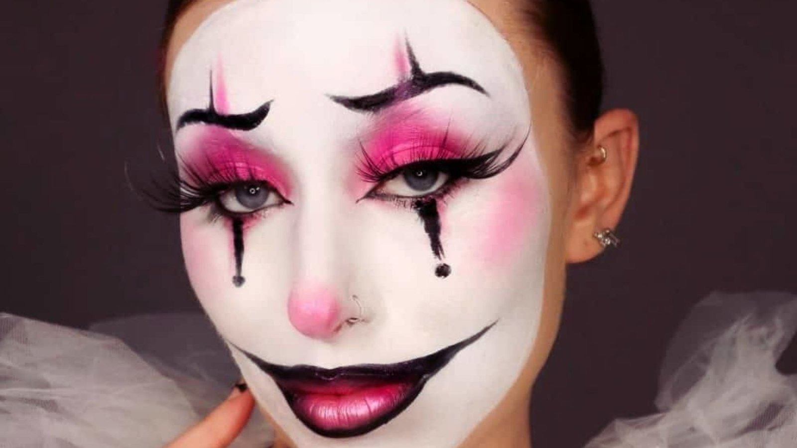 A woman with colorful clown face paint, featuring bright colors and exaggerated features, smiles playfully at the camera.
