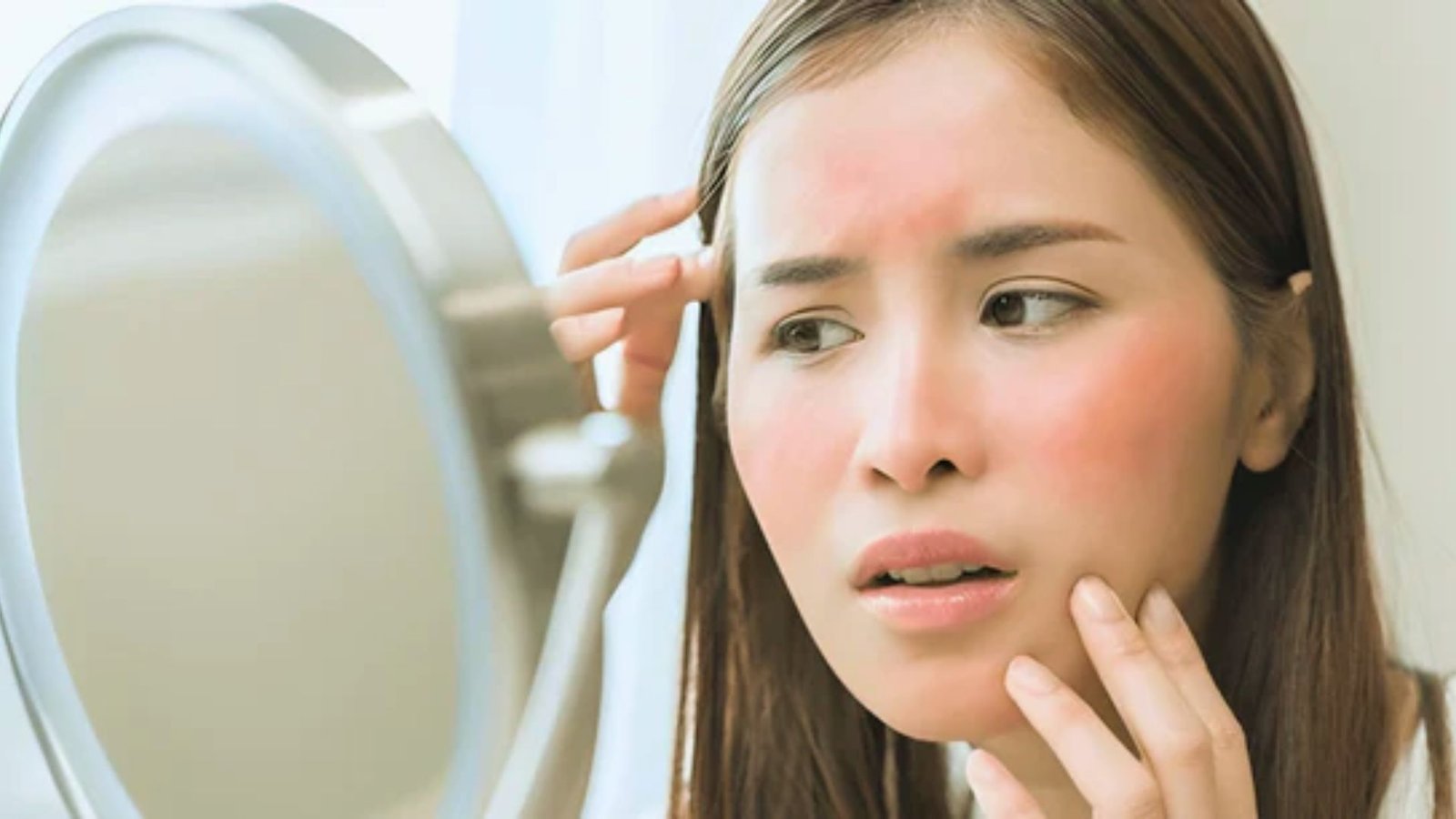 A woman gazes into a mirror, examining her skin with a thoughtful expression.
