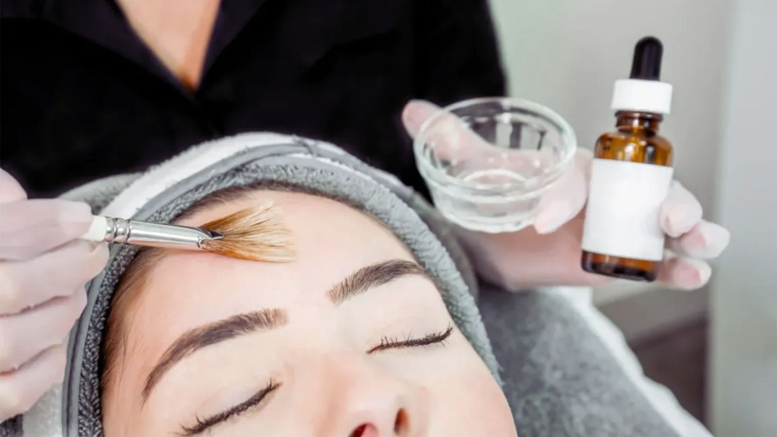  A woman having her eyebrows shaped, with a bottle of serum placed nearby on the table.
