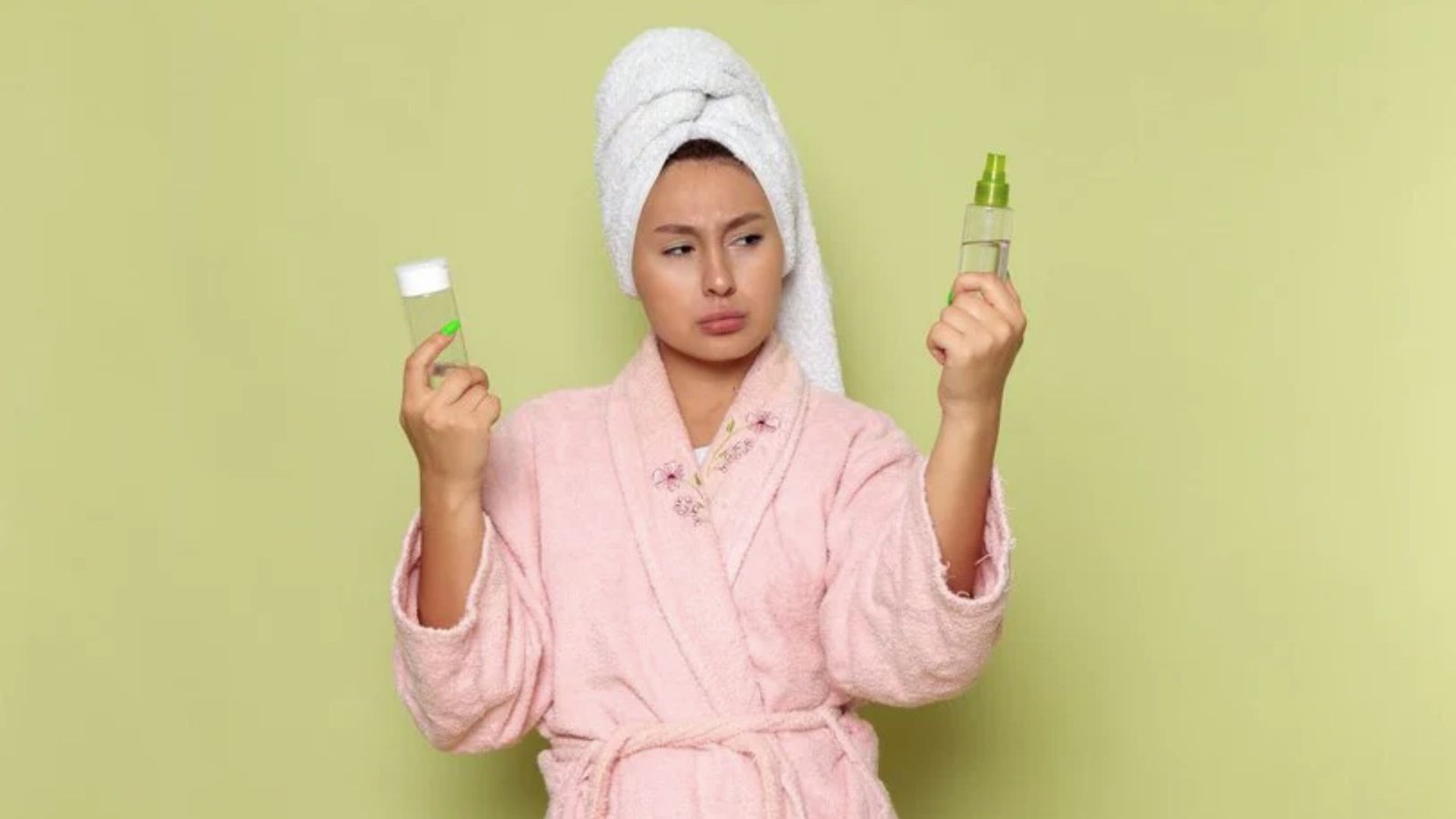 Woman in a pink robe and towel on her head looks confused, holding two skincare bottles against a light green background, conveying indecision.