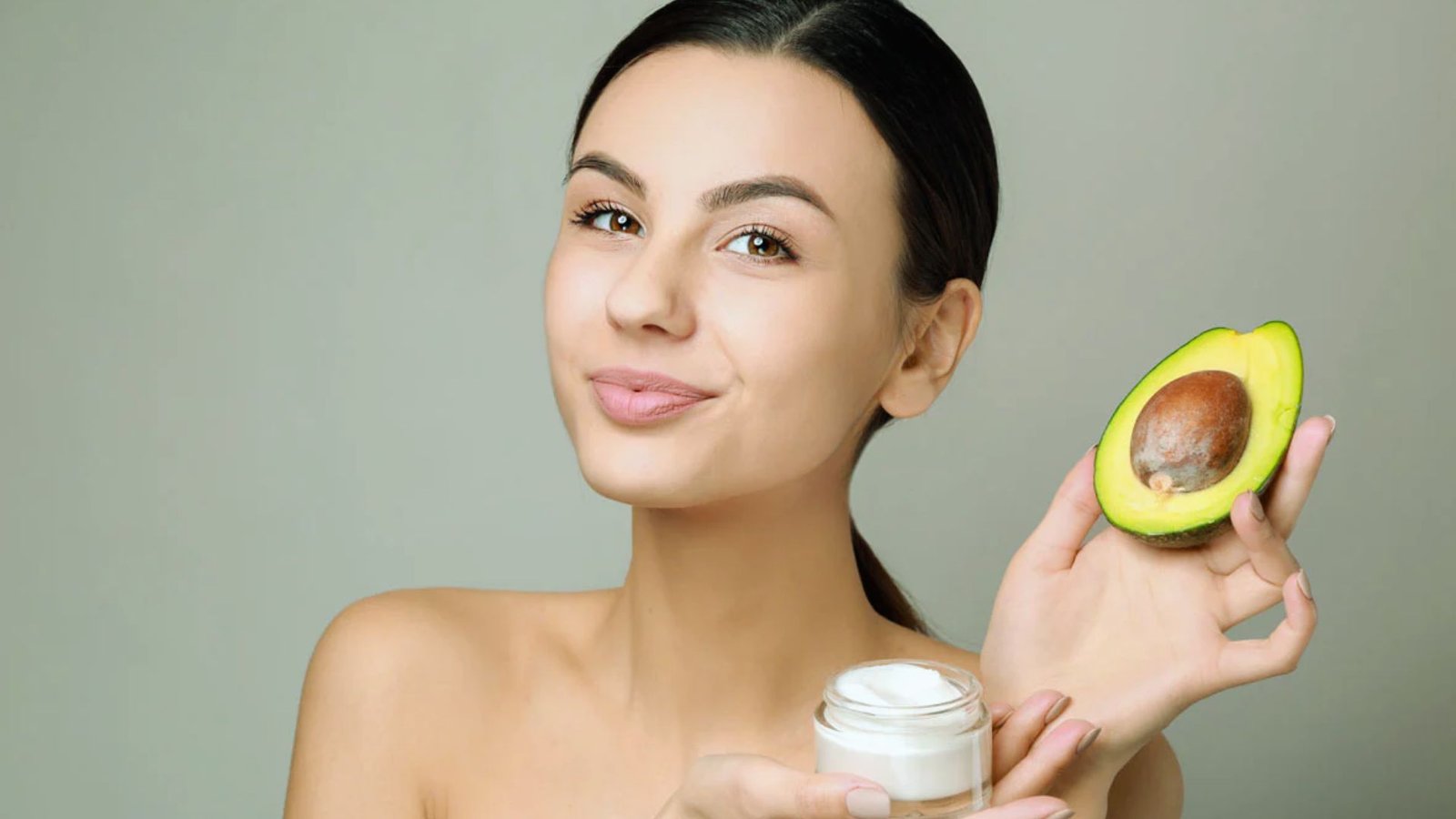 A woman with a gentle smile holds half an avocado in one hand and a small jar of cream in the other against a neutral background, conveying a sense of natural skincare.