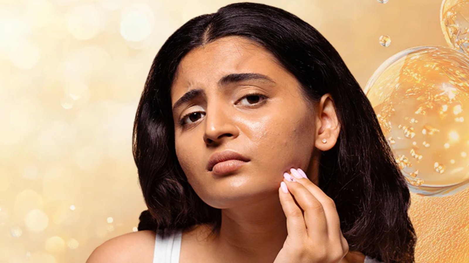 Young woman with a concerned expression touches her cheek, highlighting skin imperfections. Warm, glowing background suggests skincare focus.