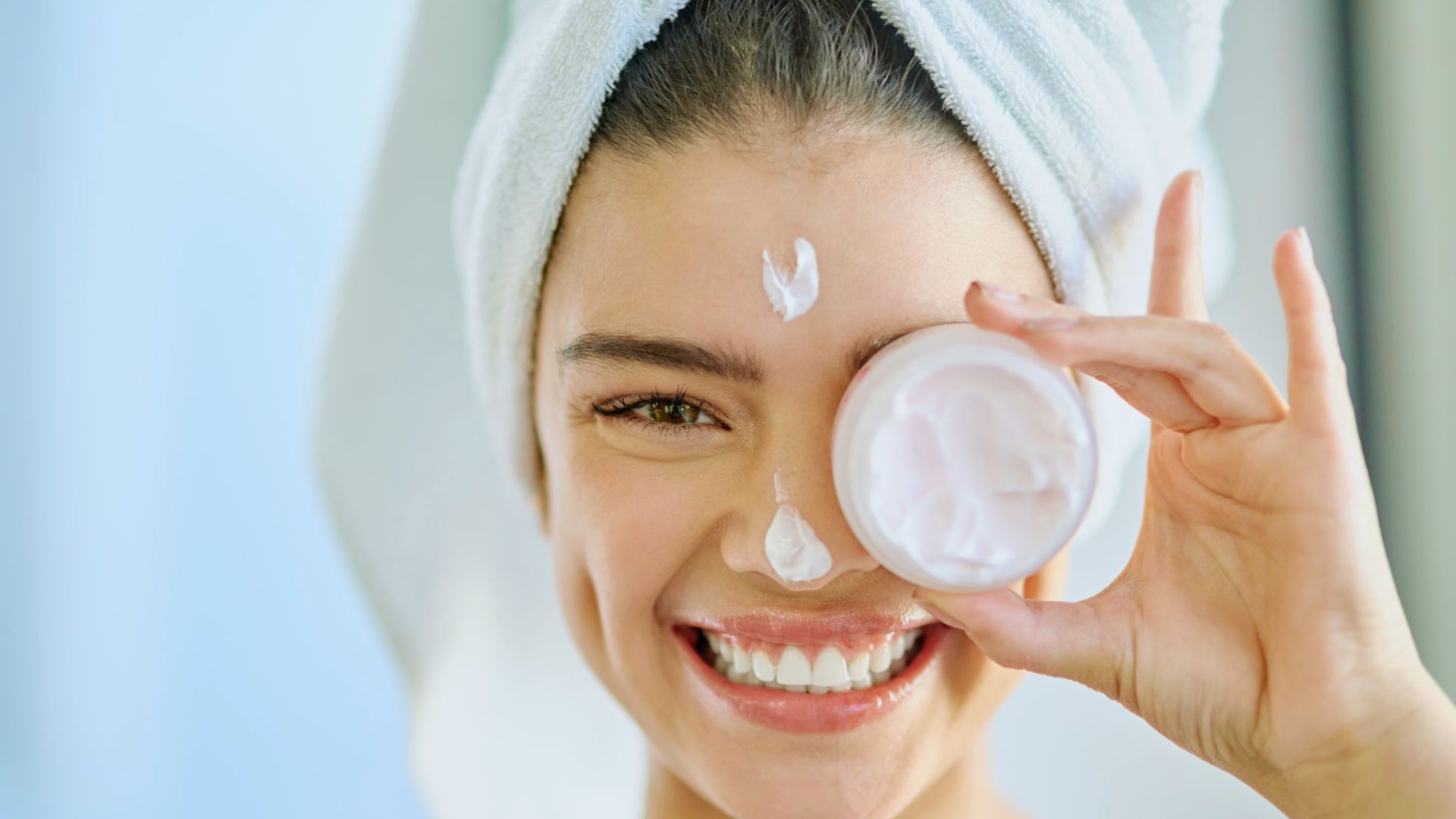 Smiling woman with a towel wrapped on her head applies face cream. She holds a jar in front of one eye. The mood is fresh and joyful.
