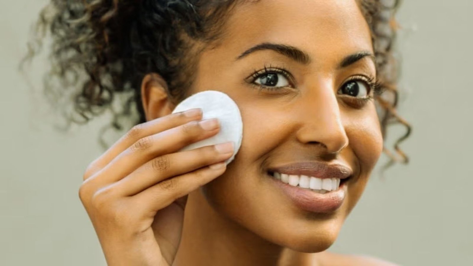 A woman with curly hair smiles as she gently applies a cotton pad to her cheek. The background is neutral, emphasizing a fresh, skincare routine.