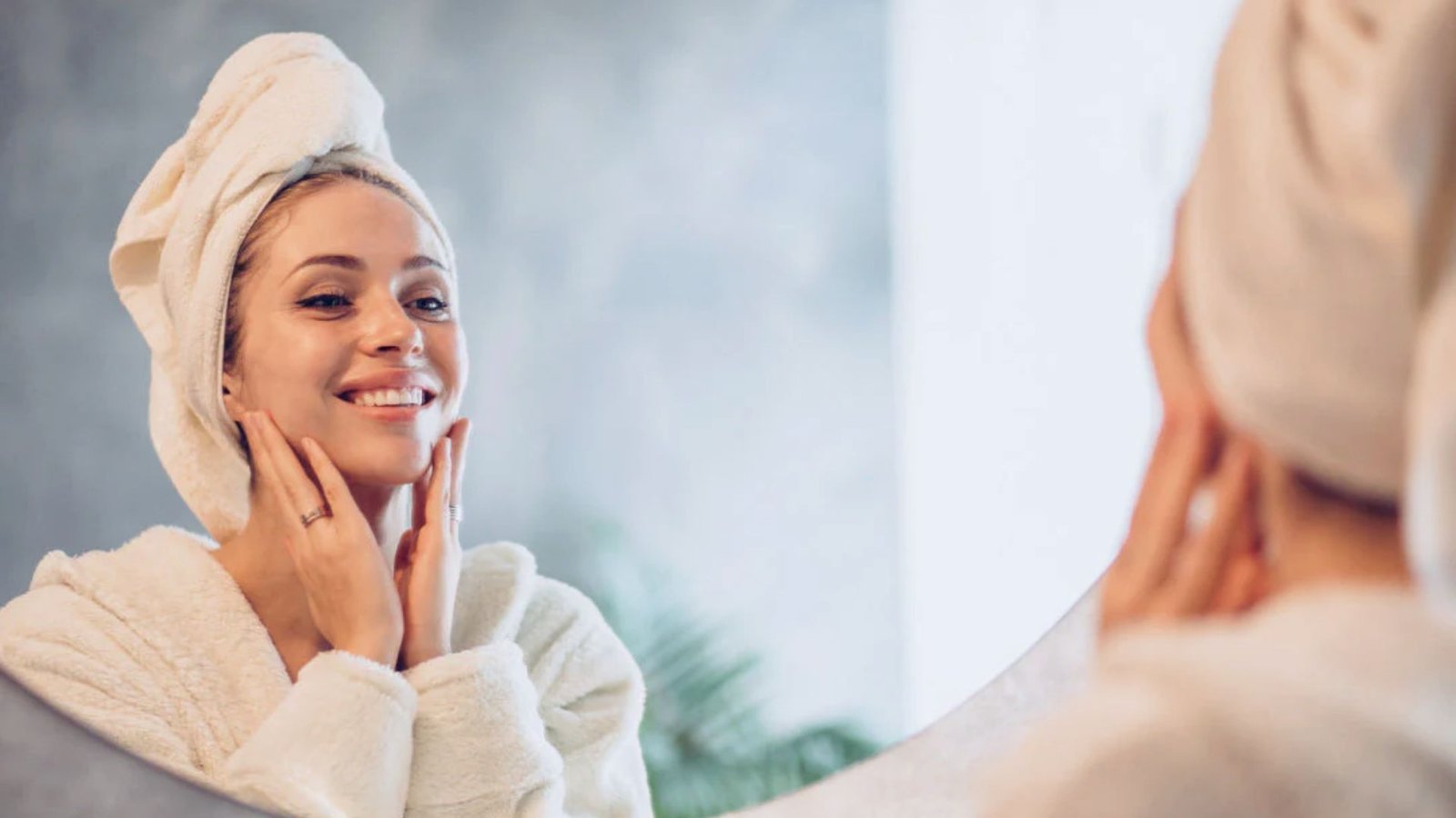 Woman with a towel on her head smiles in the mirror, gently touching her face. She's in a white robe, conveying a relaxed, content mood in a spa-like setting.