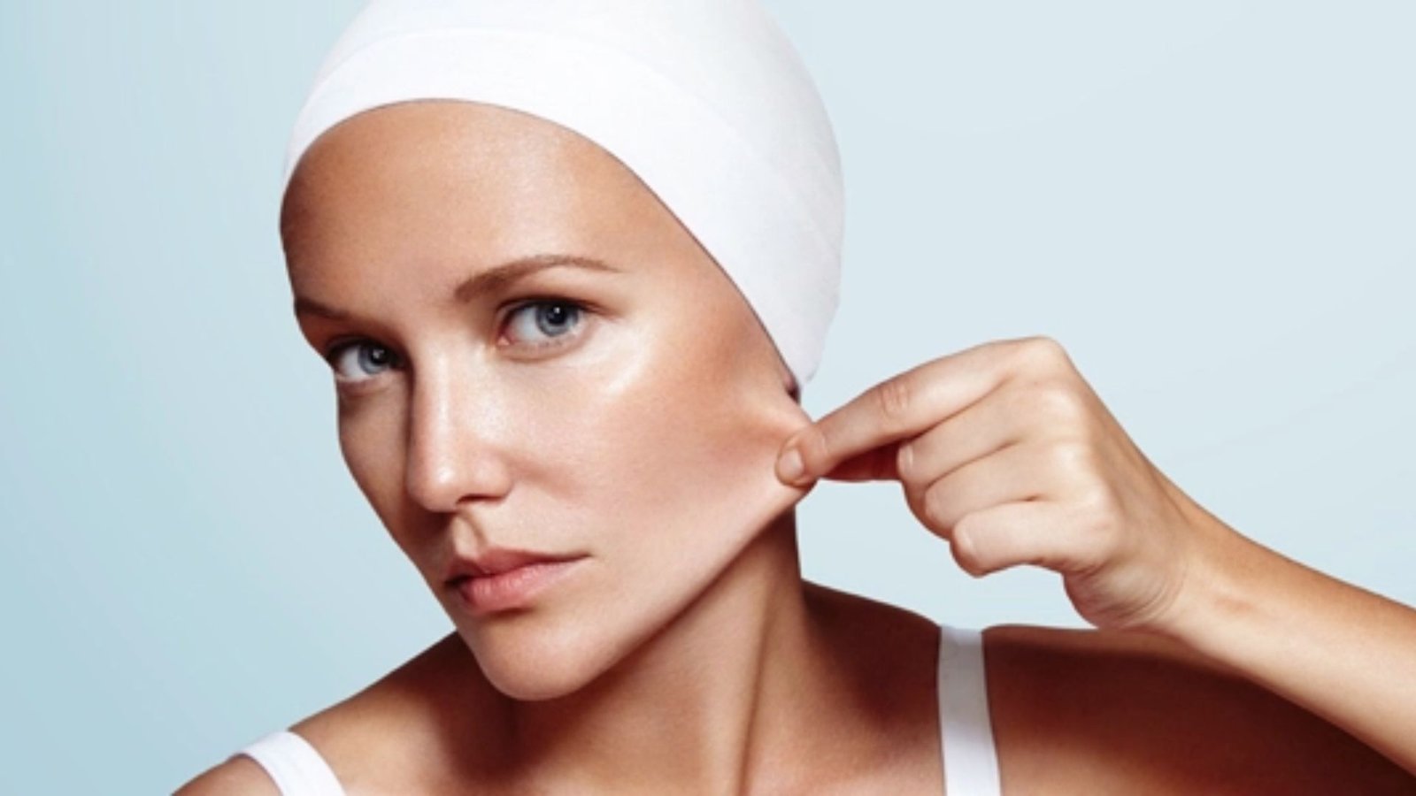 A person wearing a white head wrap pulls the skin on their cheek, demonstrating skin elasticity. The background is light blue, conveying a calm tone.
