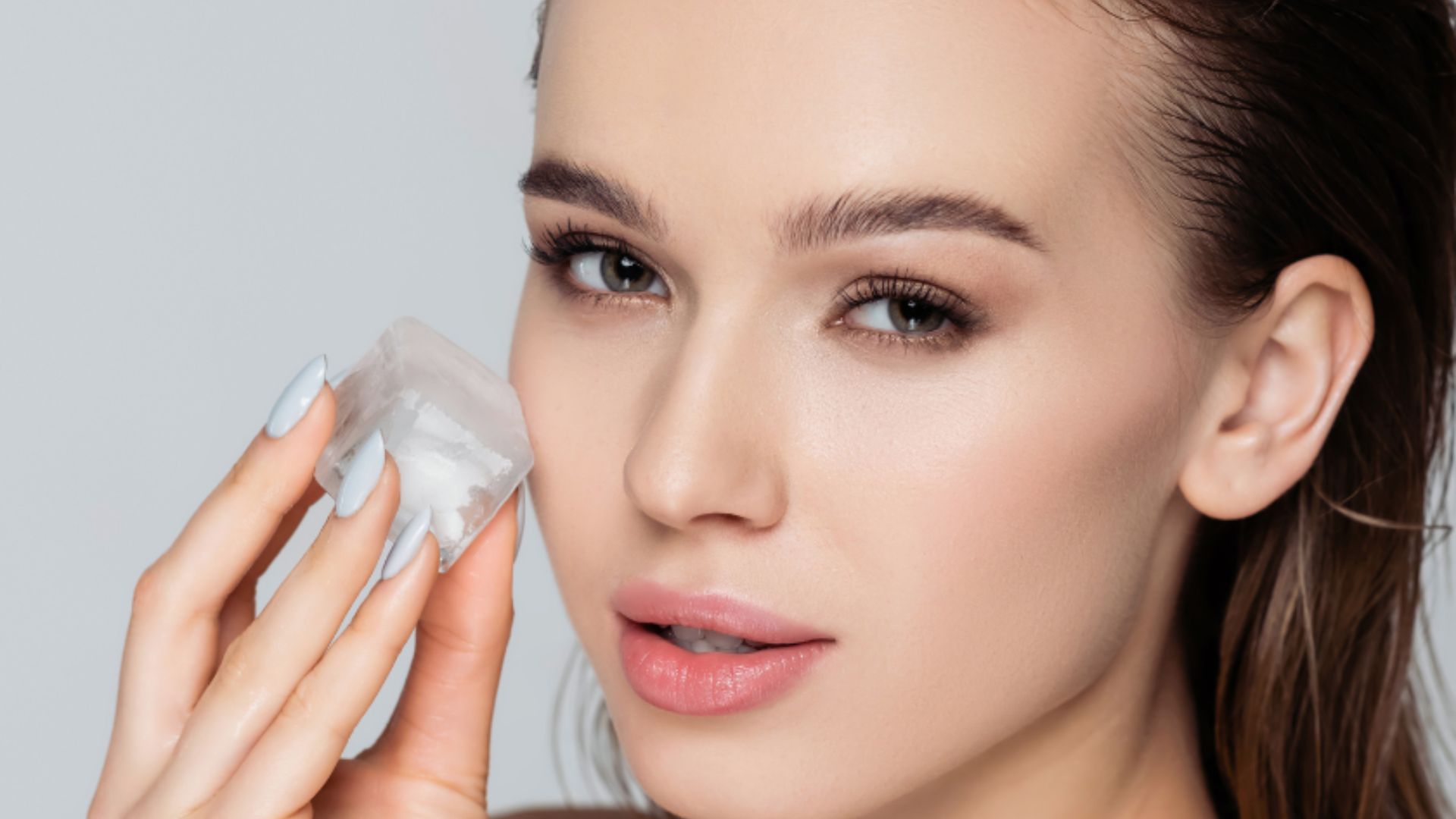 A person holds an ice cube to their cheek, showcasing a skincare routine. The serene expression and soft lighting create a fresh, rejuvenating atmosphere.