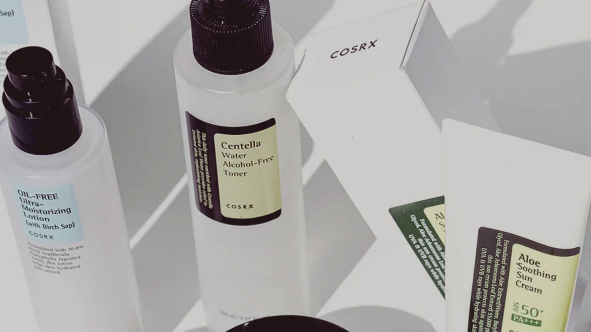 Assorted skincare products from COSRX are arranged on a white surface, including a toner and sun cream. The scene conveys a clean, minimalist aesthetic.