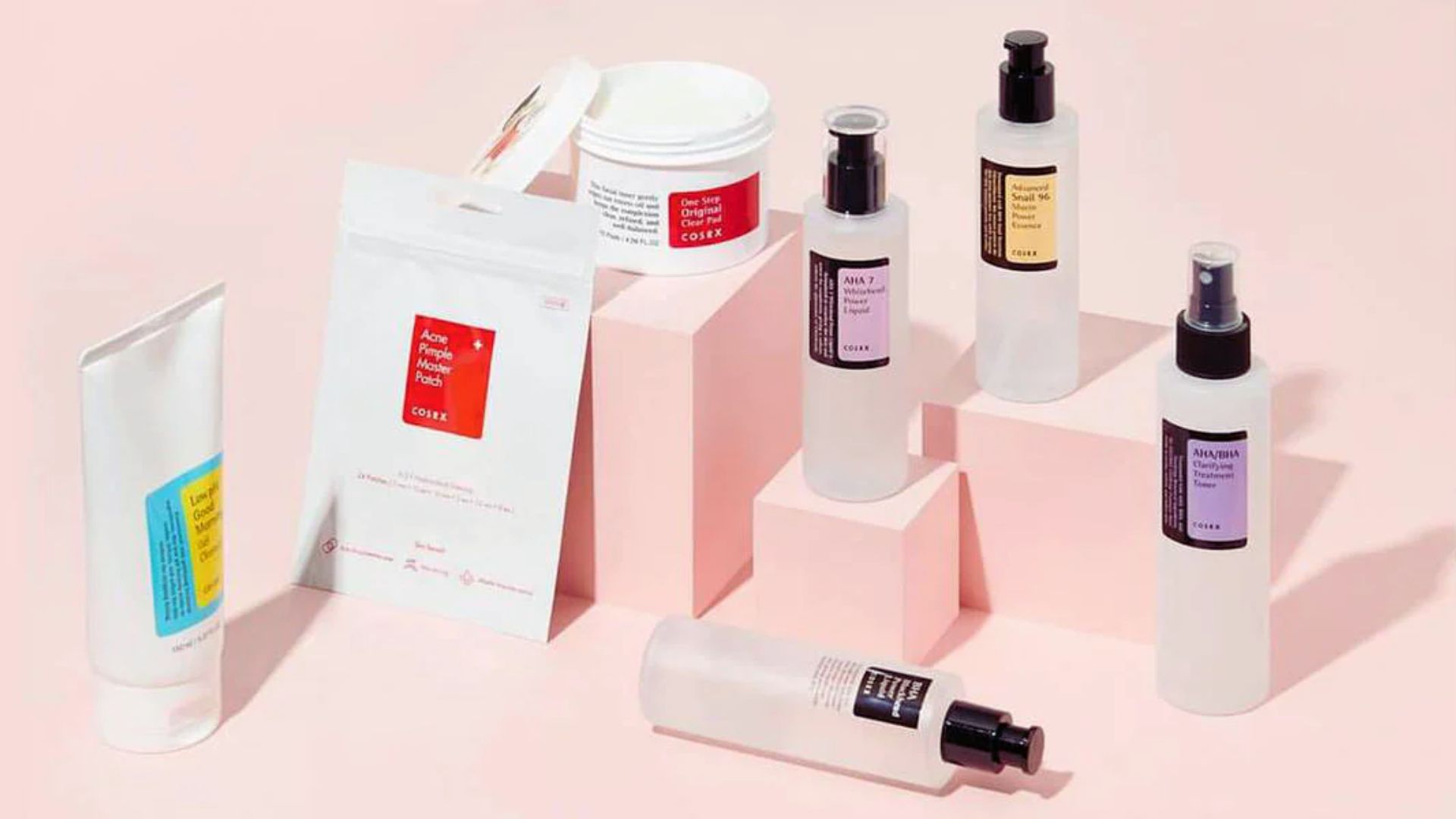 A collection of skincare products displayed on pink geometric blocks against a matching background. Various bottles and packaging create a minimalist, clean aesthetic.
