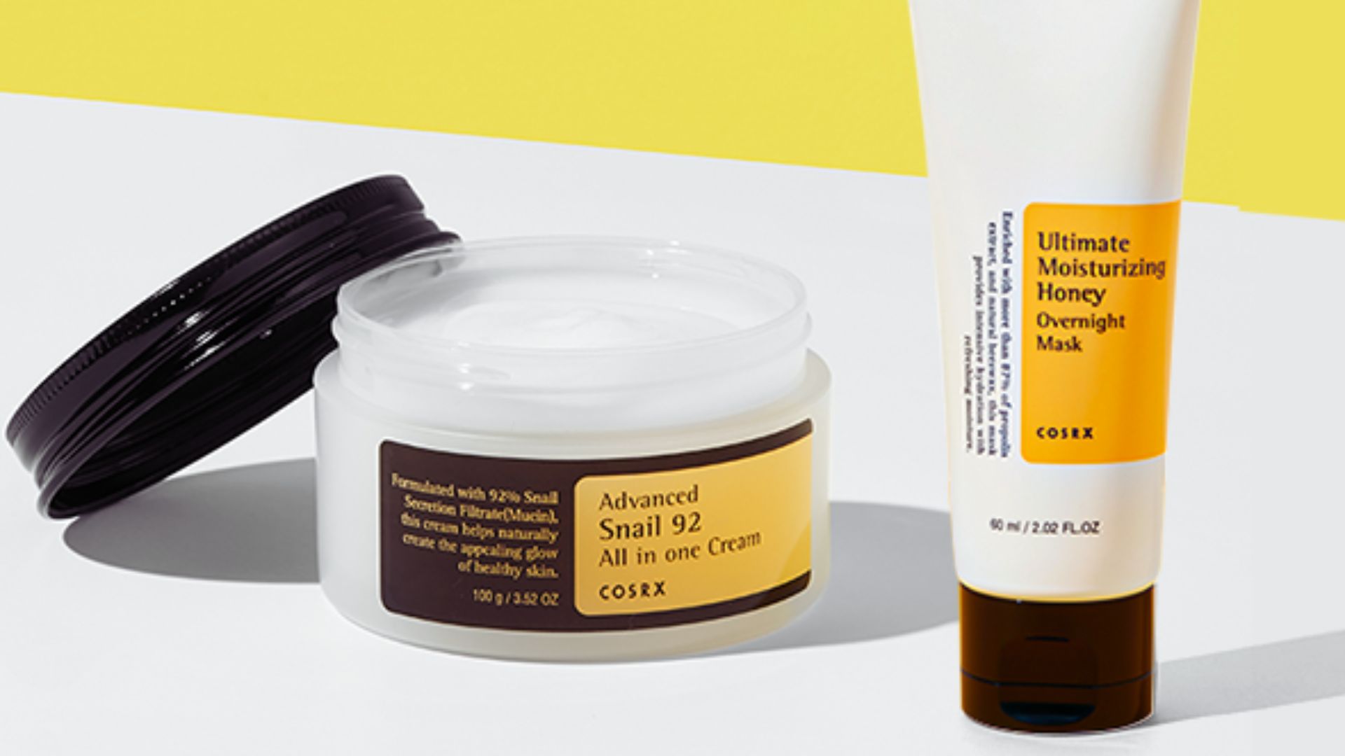 Open jar of COSRX Advanced Snail 92 Cream with a black lid beside a tube of Ultimate Moisturizing Honey Overnight Mask against a neutral backdrop.