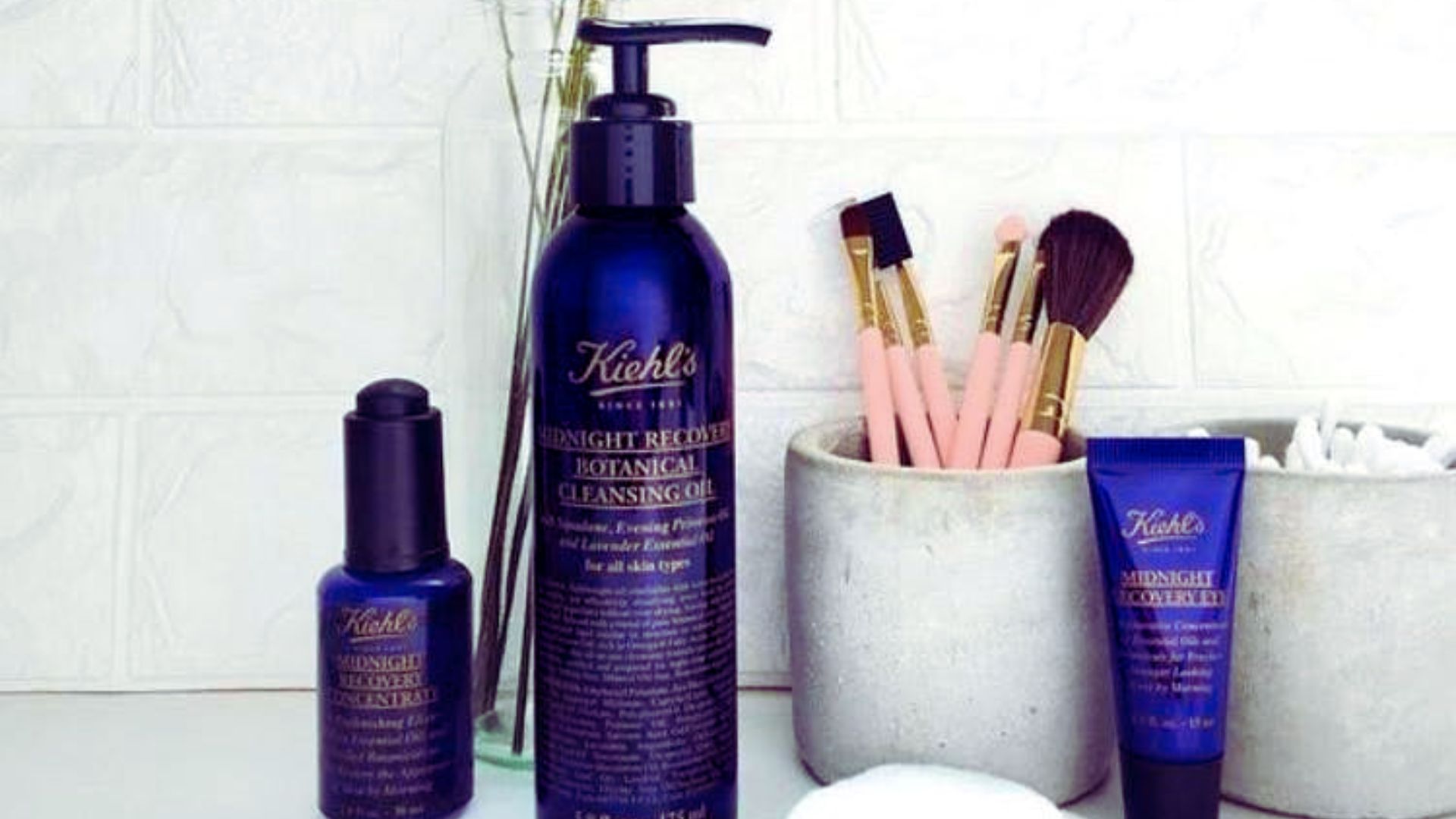 Dark blue Kiehl's skincare bottles are on a bathroom counter. Nearby, makeup brushes sit in a concrete holder with cotton pads, creating a serene, spa-like atmosphere.