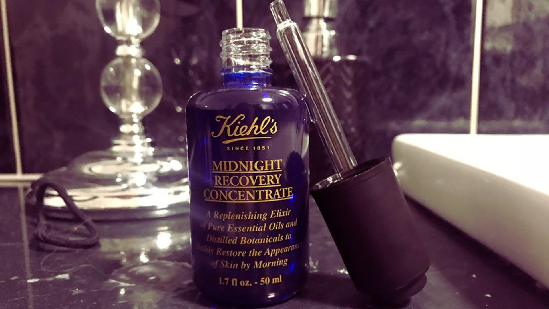 A blue bottle of Kiehl's Midnight Recovery Concentrate with a dropper rests on a bathroom counter, surrounded by elegant decor, evoking a sense of luxury.