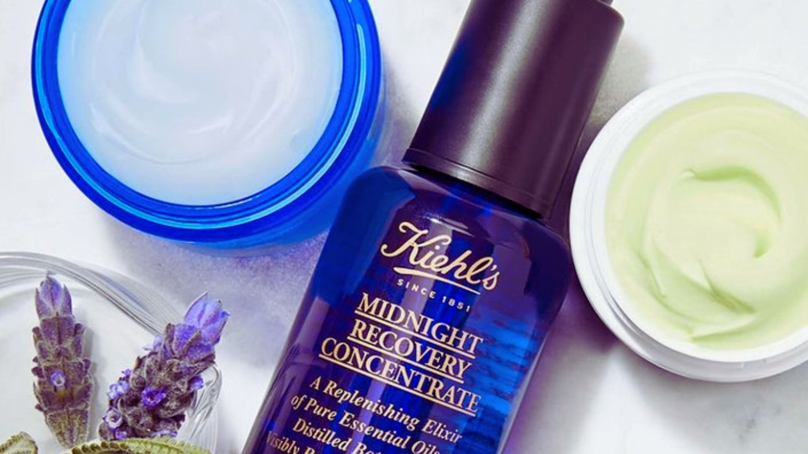 A dark blue Kiehl's Midnight Recovery Concentrate bottle is surrounded by a blue-lidded cream, a green-swirled moisturizer, and fresh lavender sprigs.