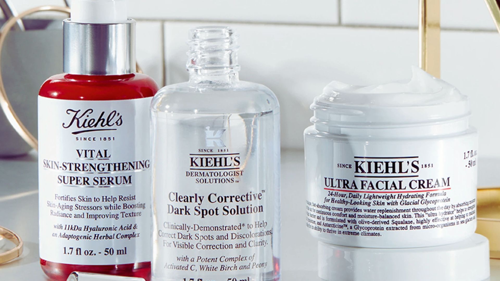 Three Kiehl's skincare products on a bathroom countertop: Vital Skin-Strengthening Super Serum, Clearly Corrective Dark Spot Solution, and Ultra Facial Cream.