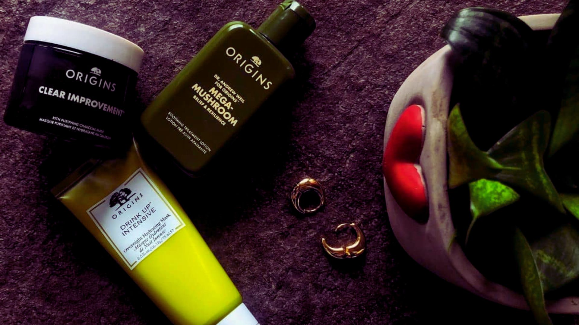 Alt text: "Flat lay of Origins skincare products on a dark surface, including Clear Improvement mask, Mega-Mushroom lotion, and Drink Up Intensive mask, alongside two small gold rings and a decorative plant in a face-shaped pot."
