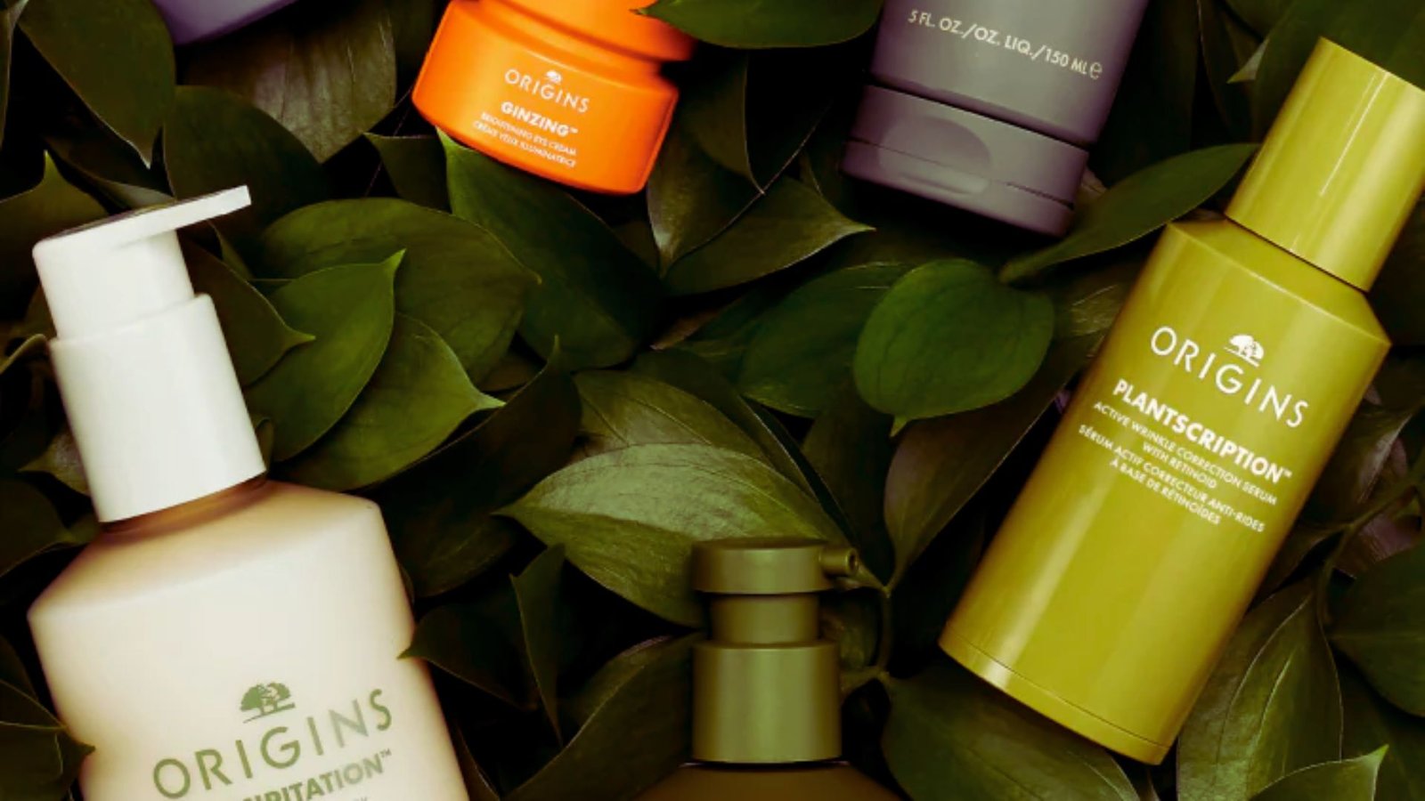 Various Origins skincare bottles in different colors are arranged on a lush bed of green leaves, suggesting a natural, organic theme.