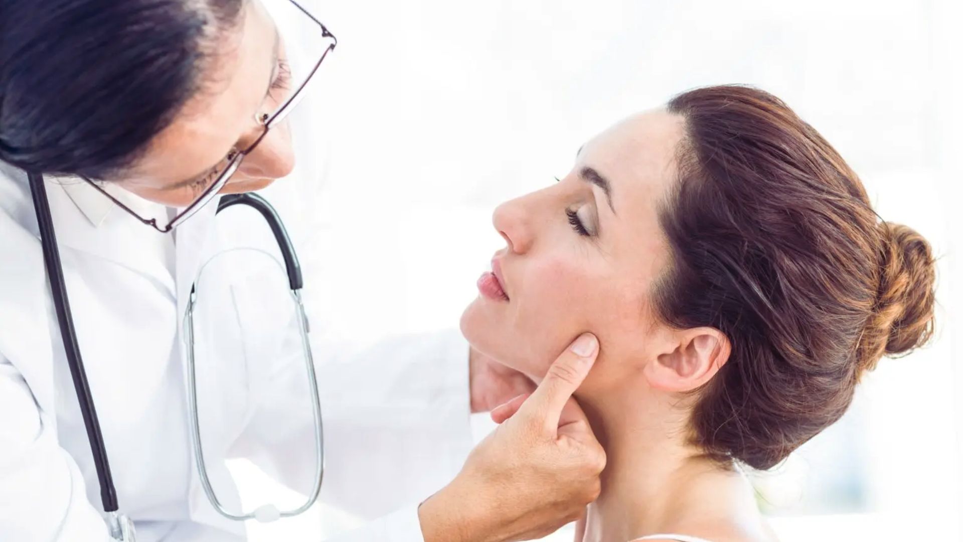 A doctor examines a woman's jawline gently with fingertips. The woman has her hair in a bun and sits with her eyes closed, conveying calmness.