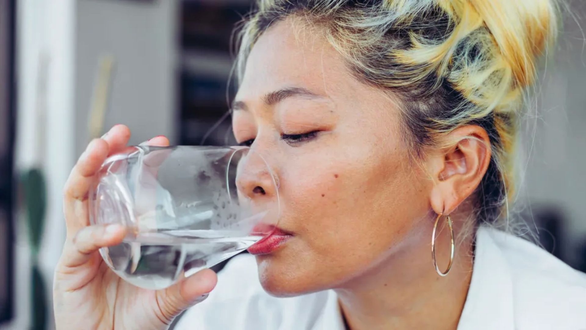 A person with blonde hair sips water from a clear glass, closed eyes suggesting refreshment and relaxation. They wear hoop earrings and a white shirt.