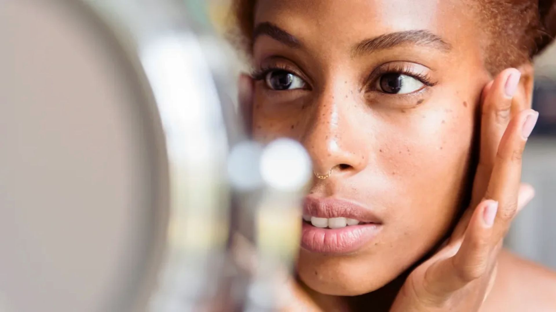 A woman with fresh, dewy skin and freckles gently touches her face while gazing into a mirror, conveying a sense of self-reflection and contemplation.