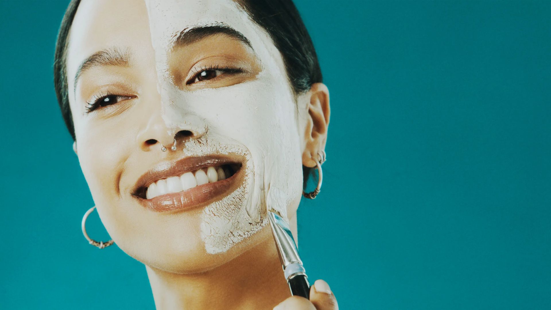Smiling woman applying white face mask with a brush on half of her face, set against a teal background. She wears hoop earrings, conveying joy and relaxation.