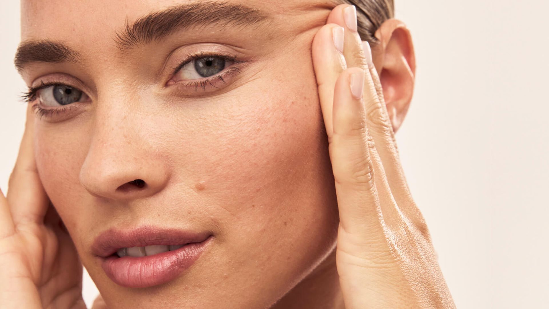 Close-up of a person gently touching their skin with both hands, showing clear, smooth complexion. The expression is calm and serene.