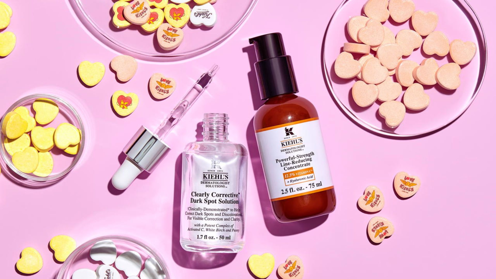 Kiehl's skincare bottles on a pink surface surrounded by colorful heart-shaped candies, creating a bright, playful atmosphere.
