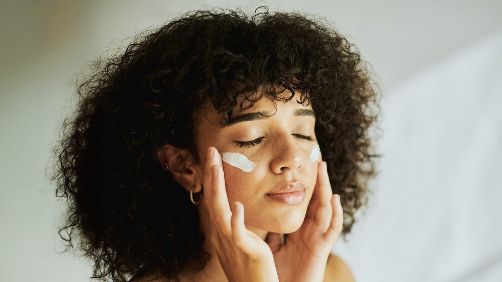 A woman with curly hair is seen applying facial cream, emphasizing her skincare routine in a cozy environment.