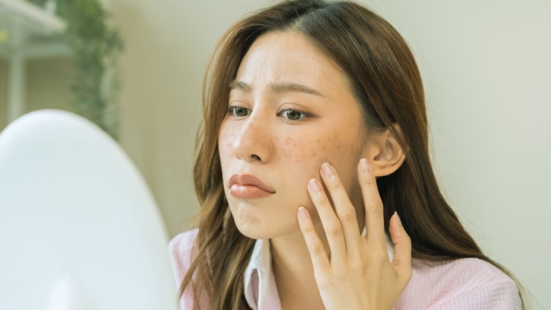 A woman with a concerned expression examines her skin in a mirror, touching her face gently. She appears focused, noticing freckles or blemishes.