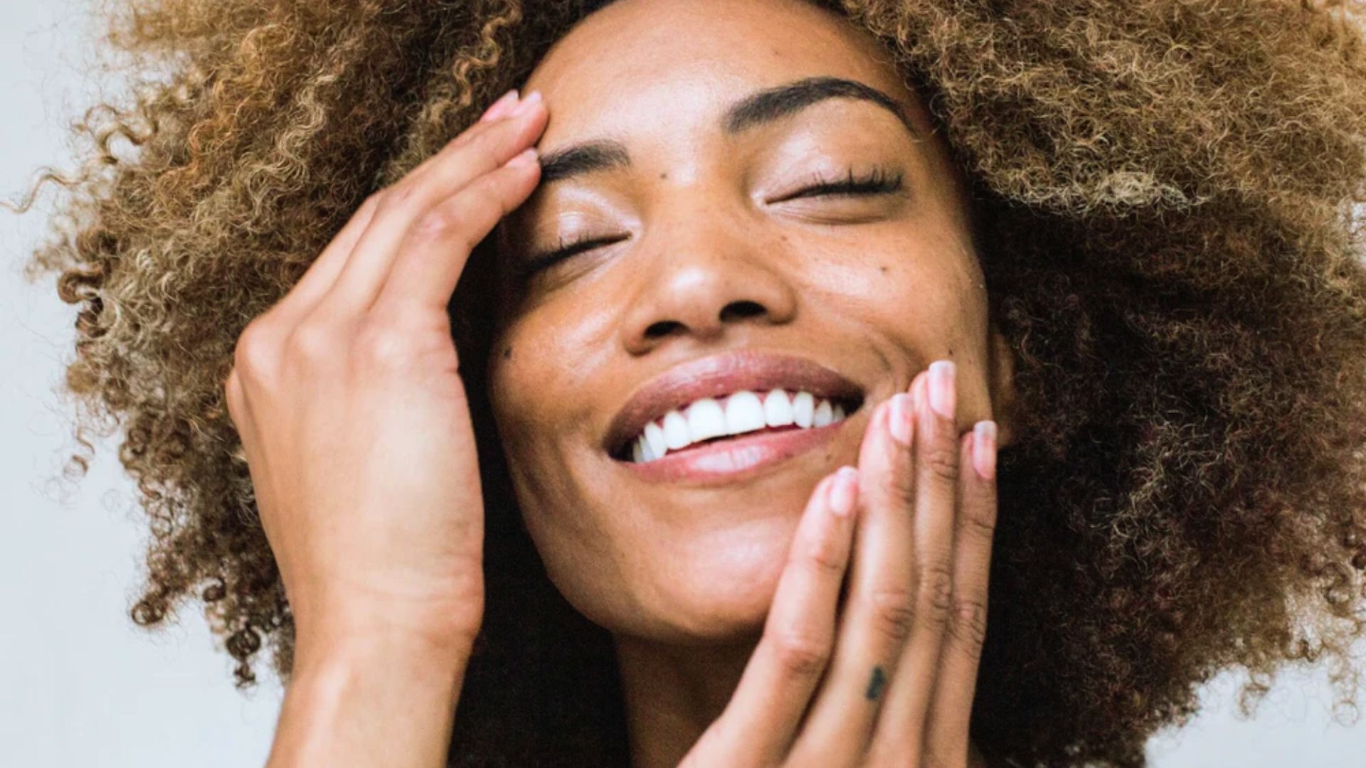A woman with eyes closed and a joyful smile, touches her face gently with both hands. Her curly hair frames her face, conveying a serene, content mood.