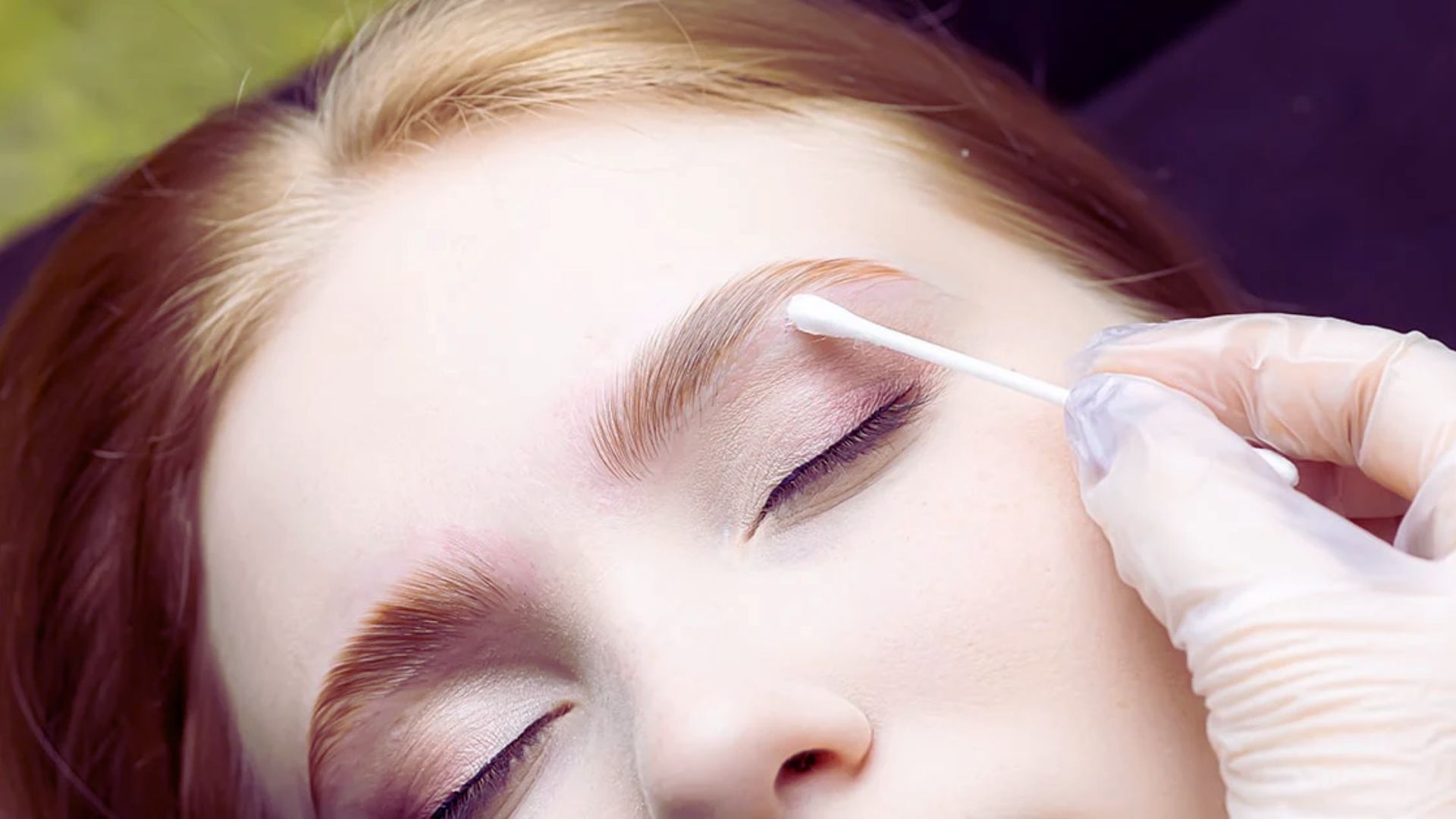 A woman sitting in a salon chair while a beautician shapes her eyebrows with tweezers.