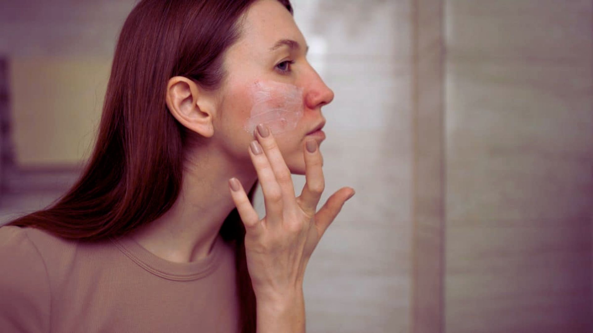 A woman applies cream to her face, focusing on skincare in a well-lit bathroom setting.