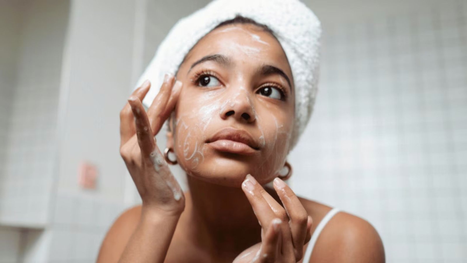 A woman wrapped in a towel applies a facial mask, focusing on her skincare routine in a bright bathroom setting.