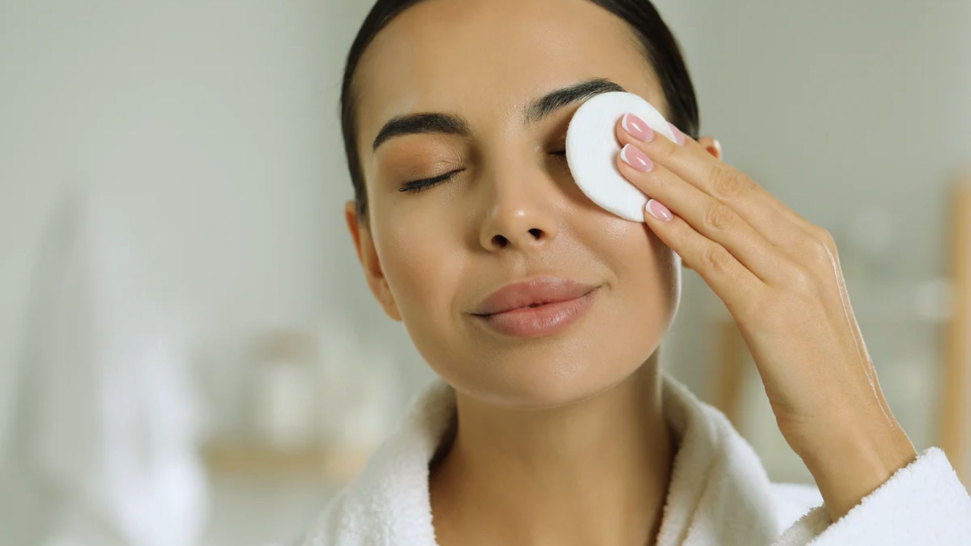 A woman in a bathrobe cleans her face with a facial sponge, focusing on her skincare routine.
