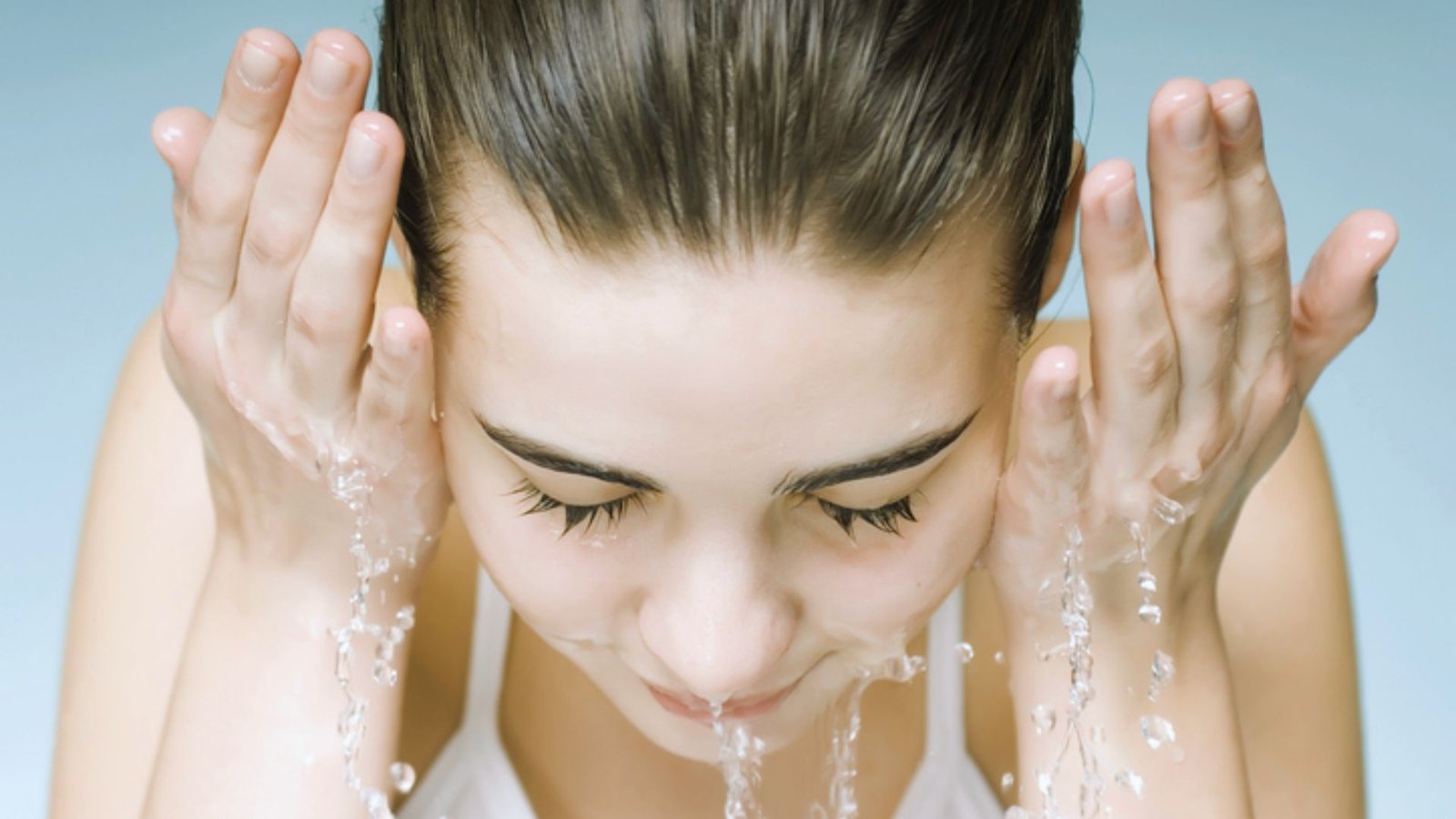 A woman is gently washing her face with water at a bathroom sink, focusing on her skincare routine.