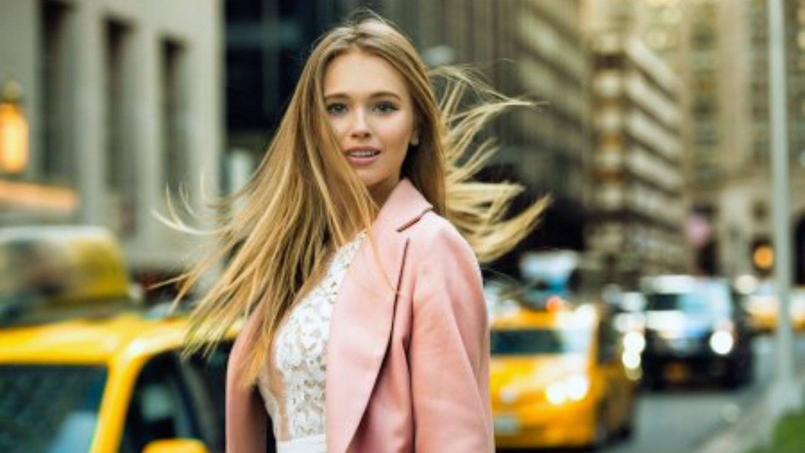 A young woman in a pink coat and white dress walks gracefully down the street, exuding confidence and style.
