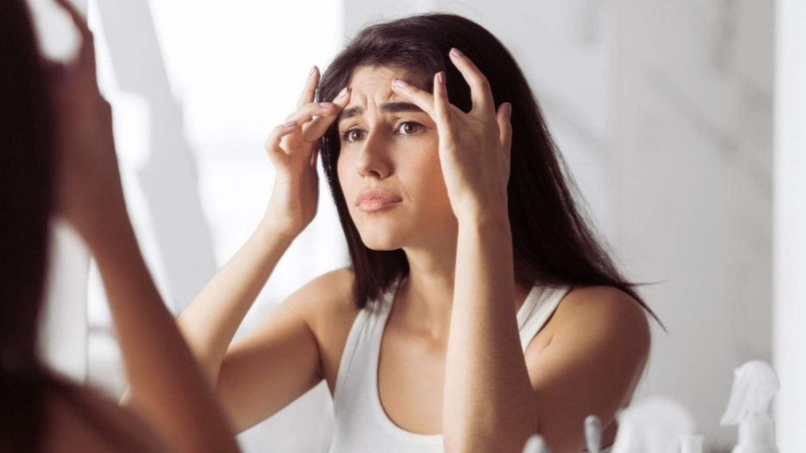 A woman gazes into a mirror, holding her head with a contemplative expression.