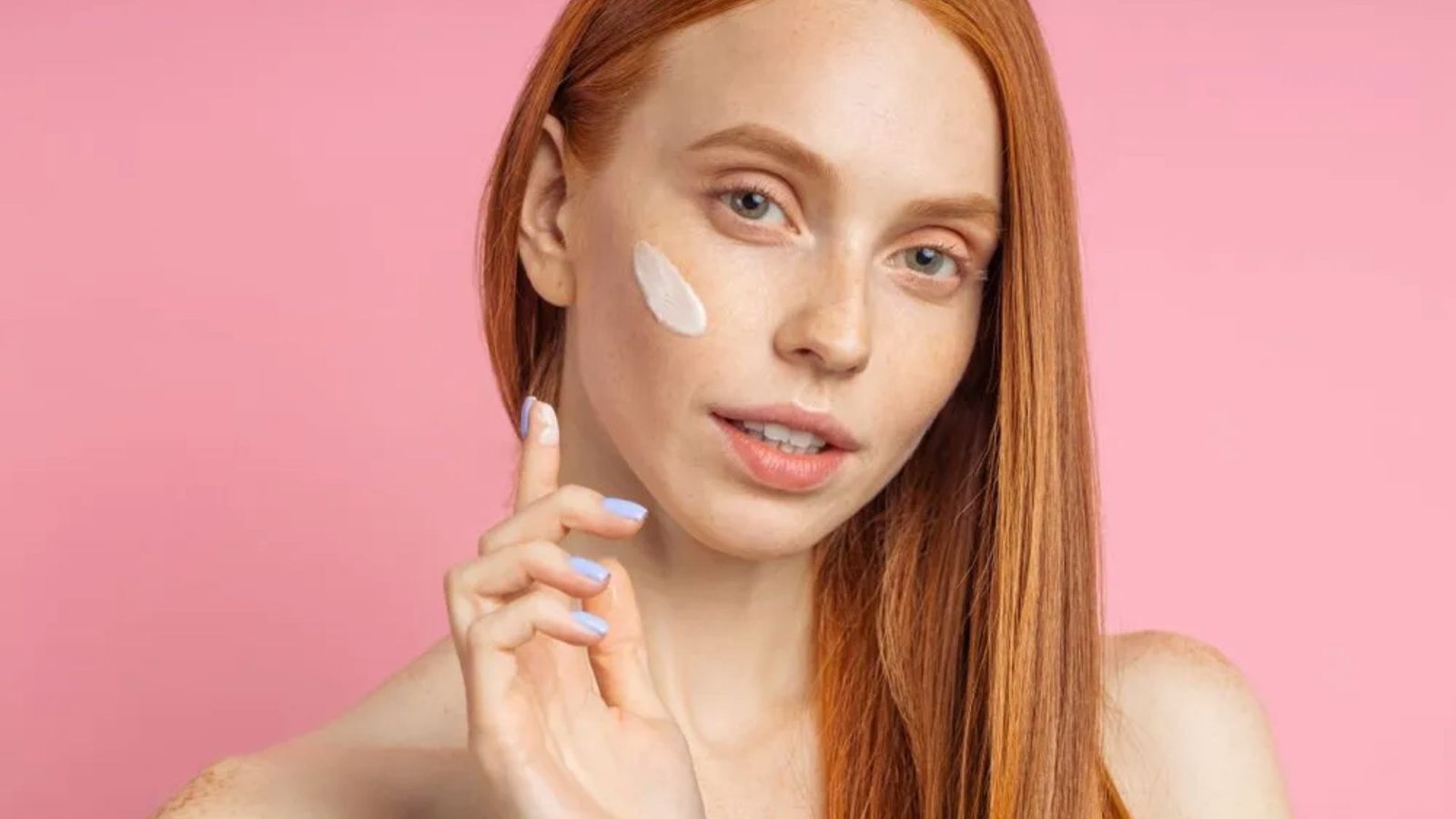 A woman with red hair applies white cream to her face, showcasing a skincare routine.
