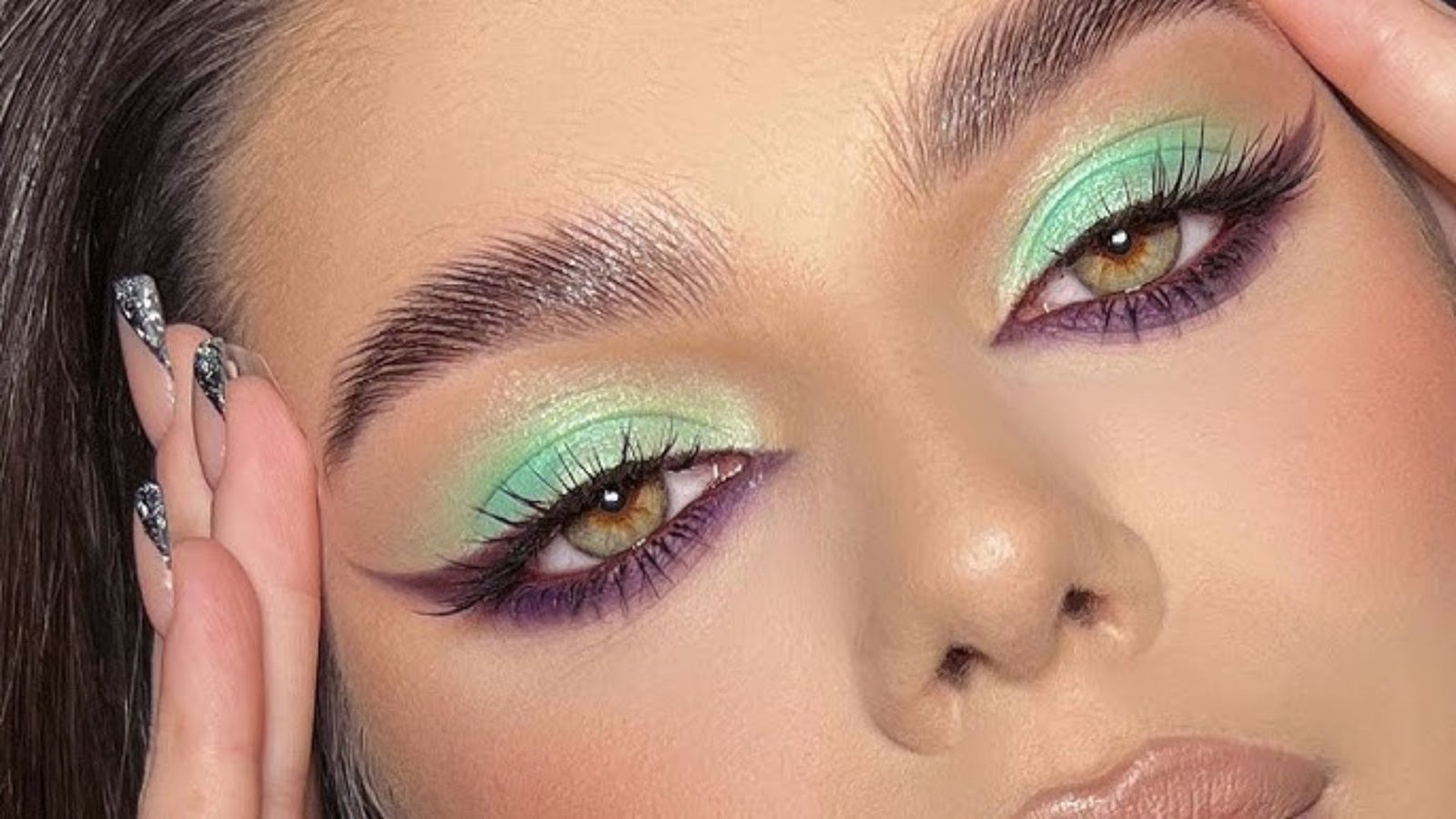 A woman with vibrant green and purple makeup artistically applied to her face, showcasing a bold and creative look.