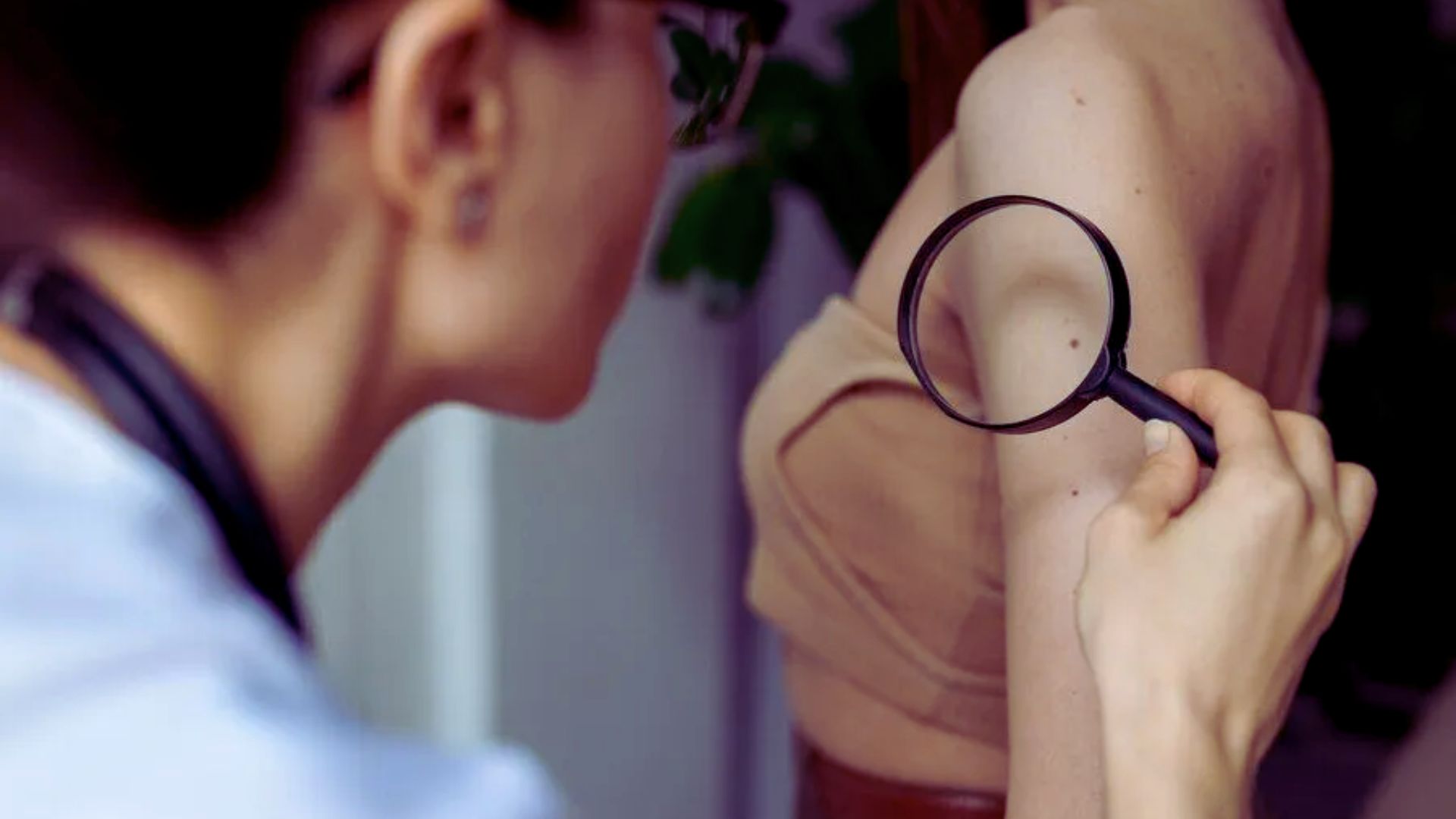 A woman examines her skin with a magnifying glass, checking for a mole on her arm.