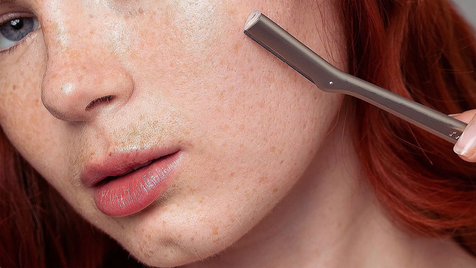 A woman with red hair shaves her face with a razor, focusing intently on her grooming routine.