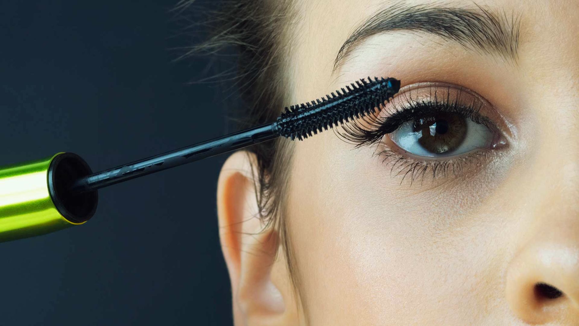 A woman applies mascara to her eyelashes using a mascara brush, focusing intently on her makeup application.