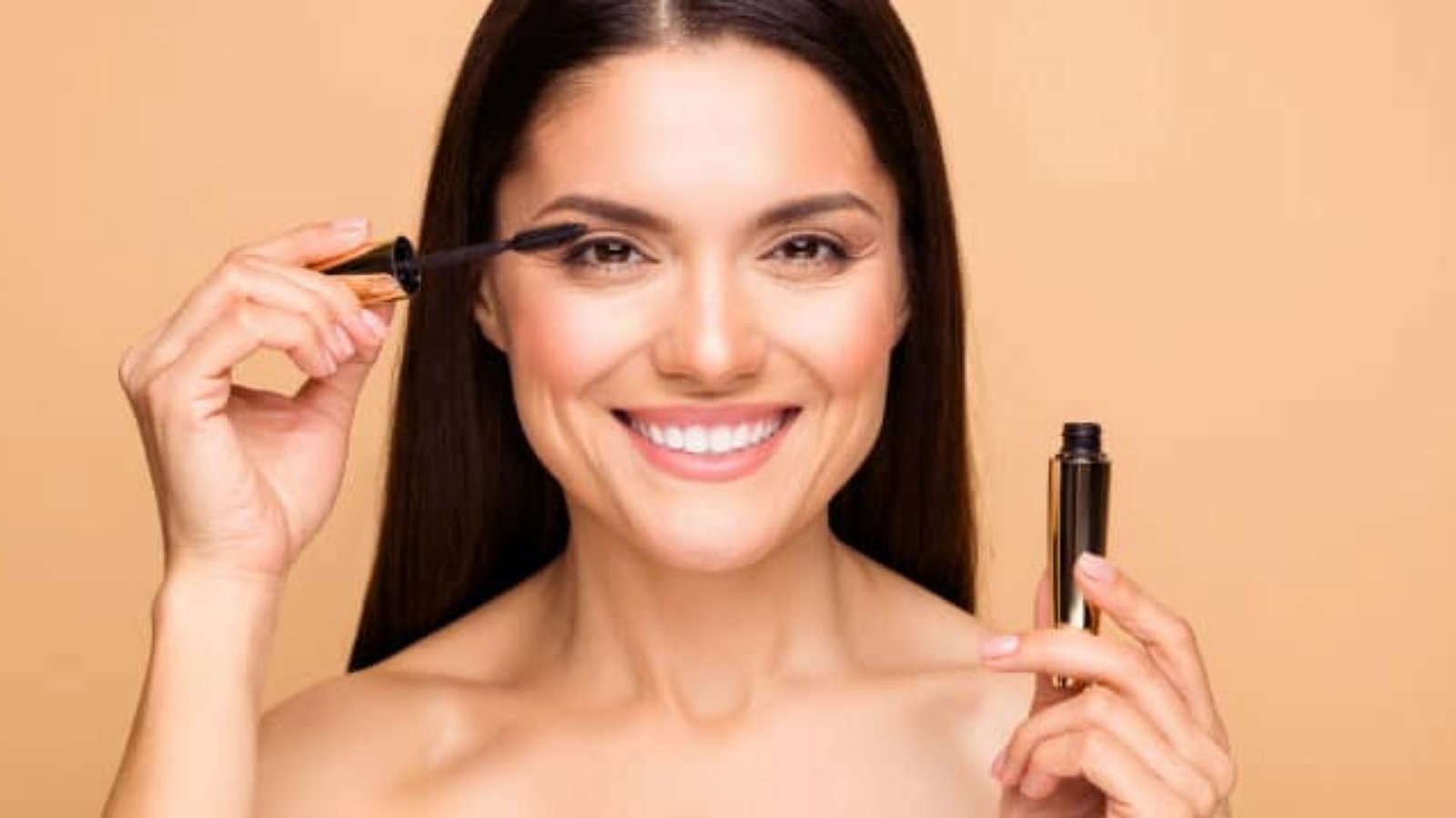 A smiling woman holds a mascara brush, showcasing her enthusiasm for makeup application.