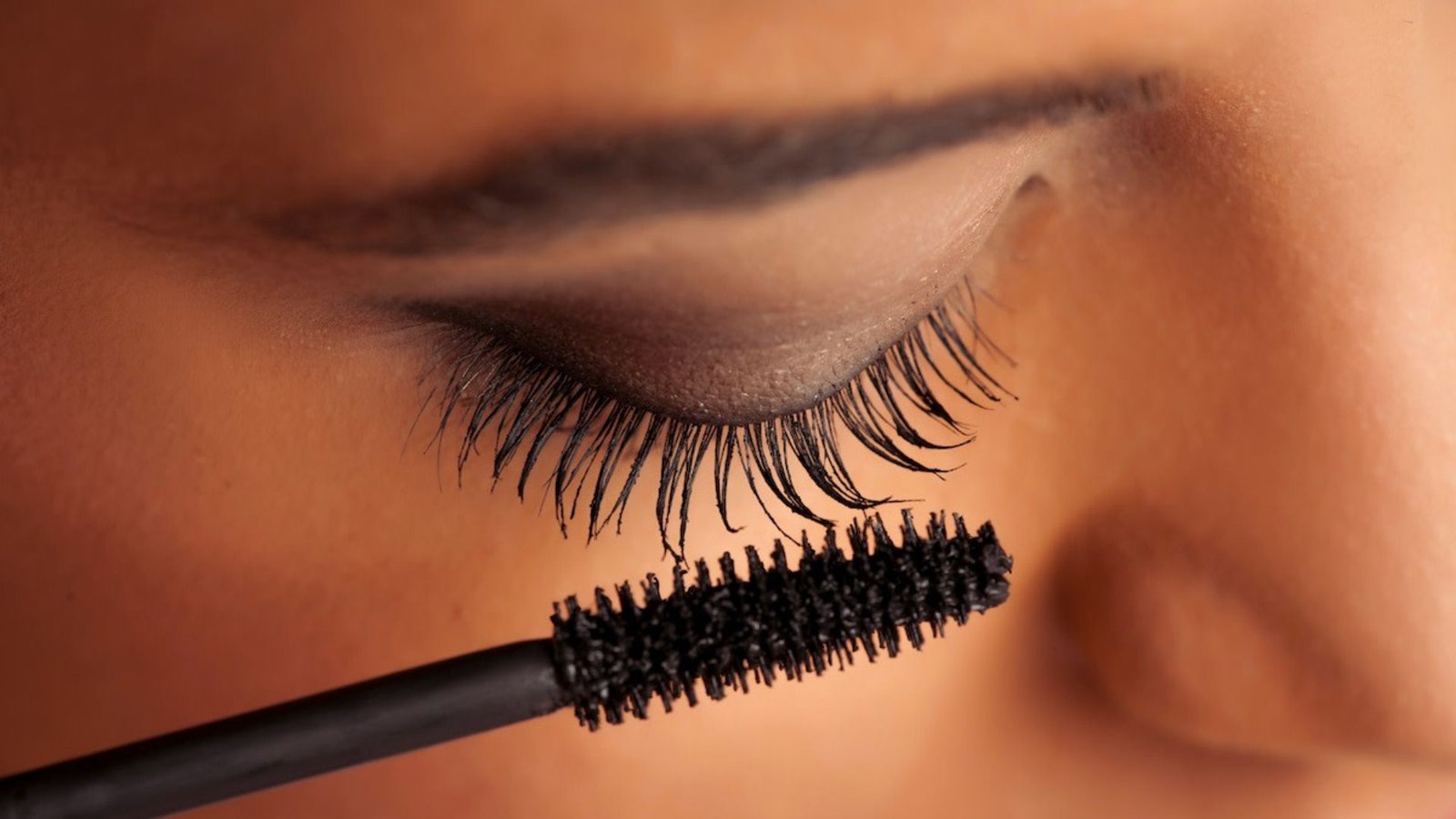 A woman applies mascara to her eyelashes using a mascara brush, focusing intently on her makeup application.